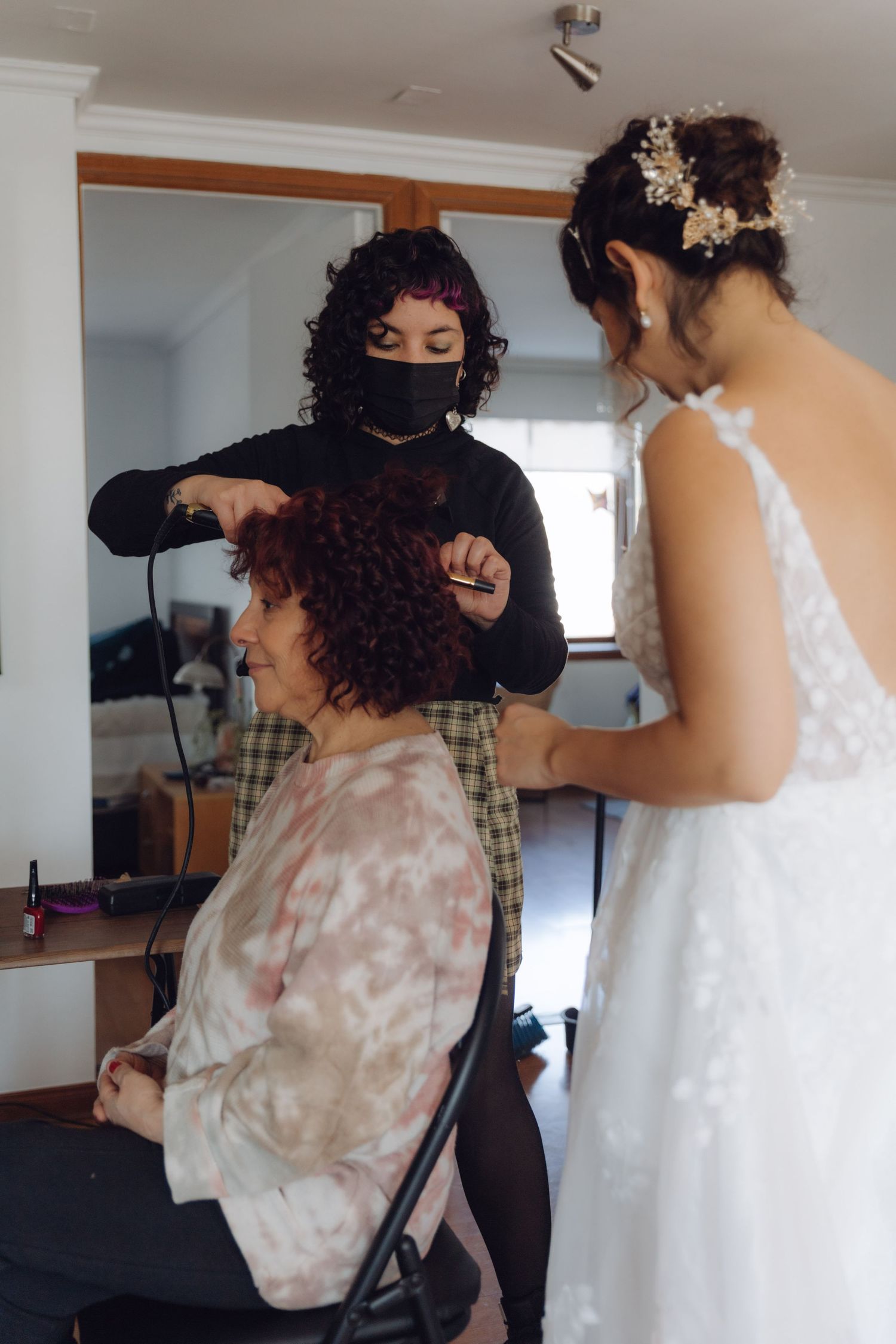 Bride in white wedding dress assisting hairstylist with guest hair during wedding day preparations.