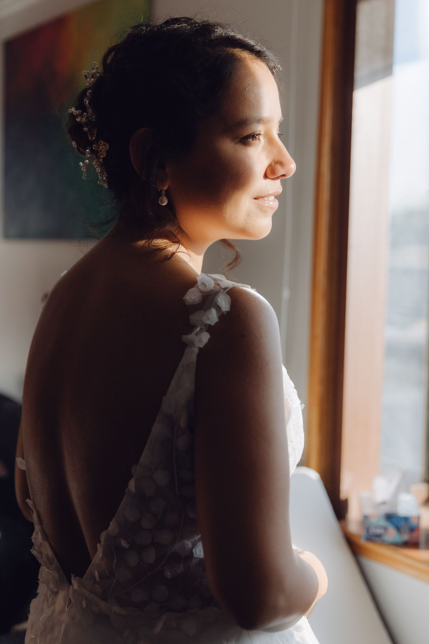 Bride in backless white wedding gown gazing out window in beautiful natural light during preparations.