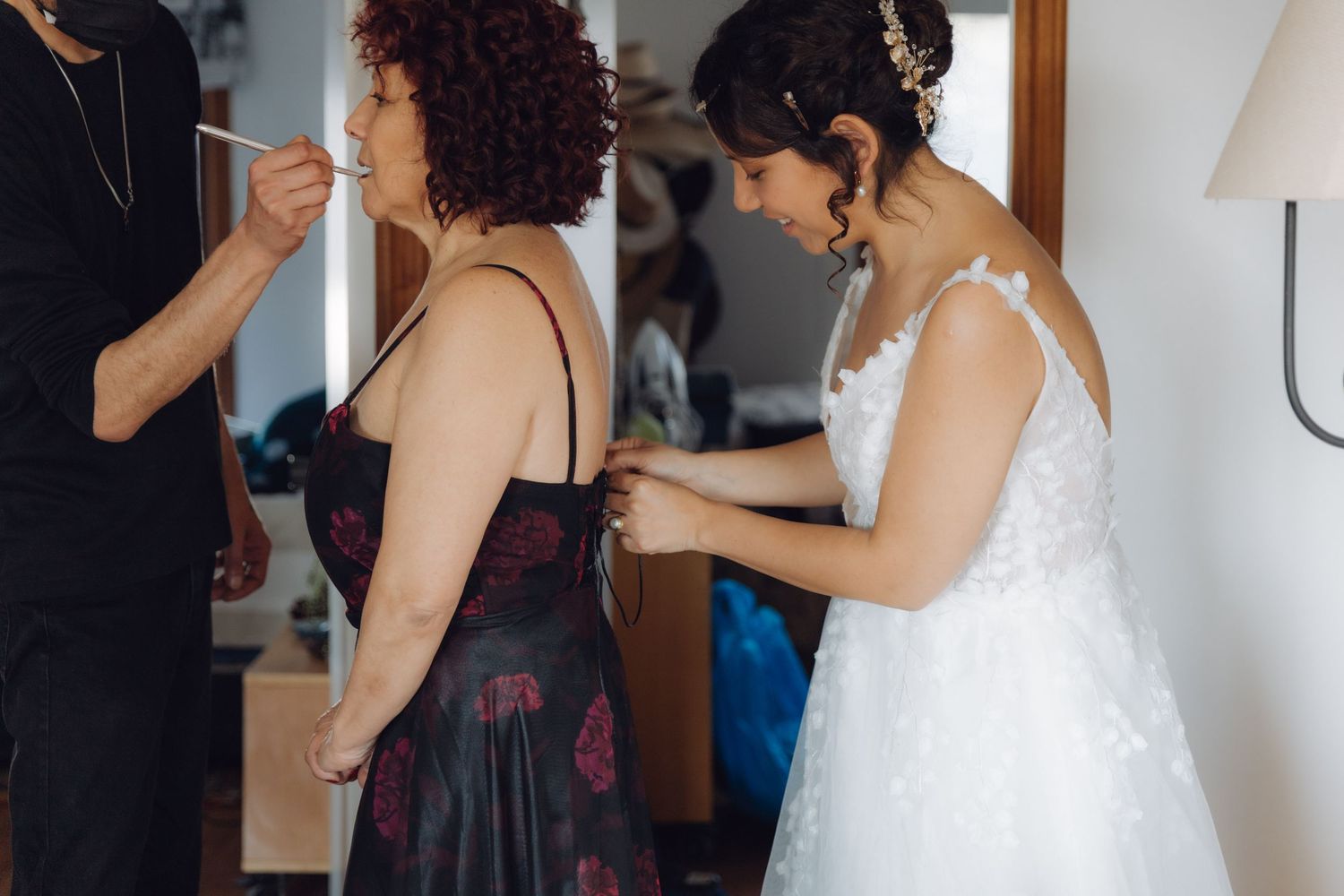 Bride in white lace gown helping fasten guest dress during wedding day getting ready preparations.