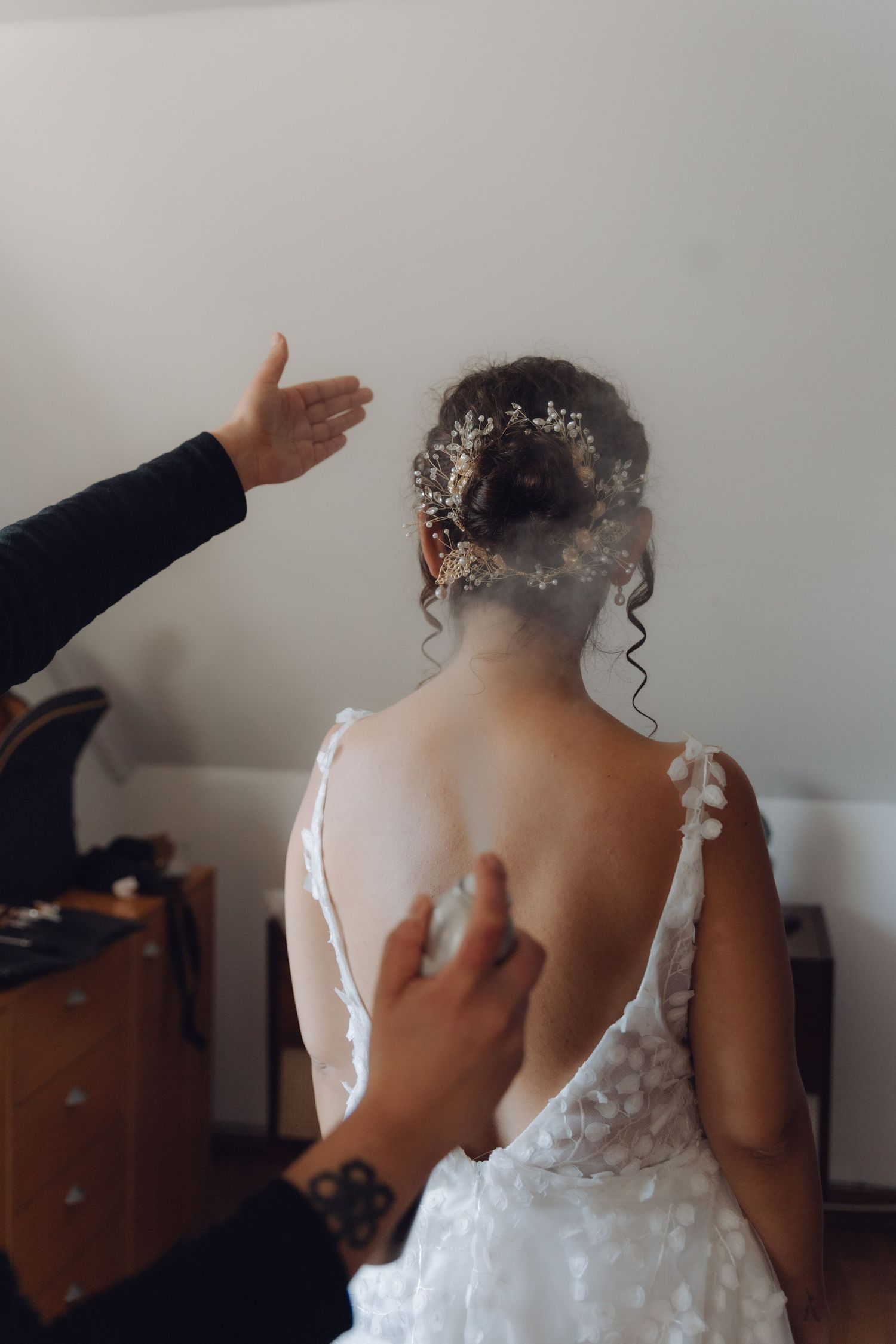 Bride getting ready as hands help style her elegant low-back white wedding dress and floral hair accessory.