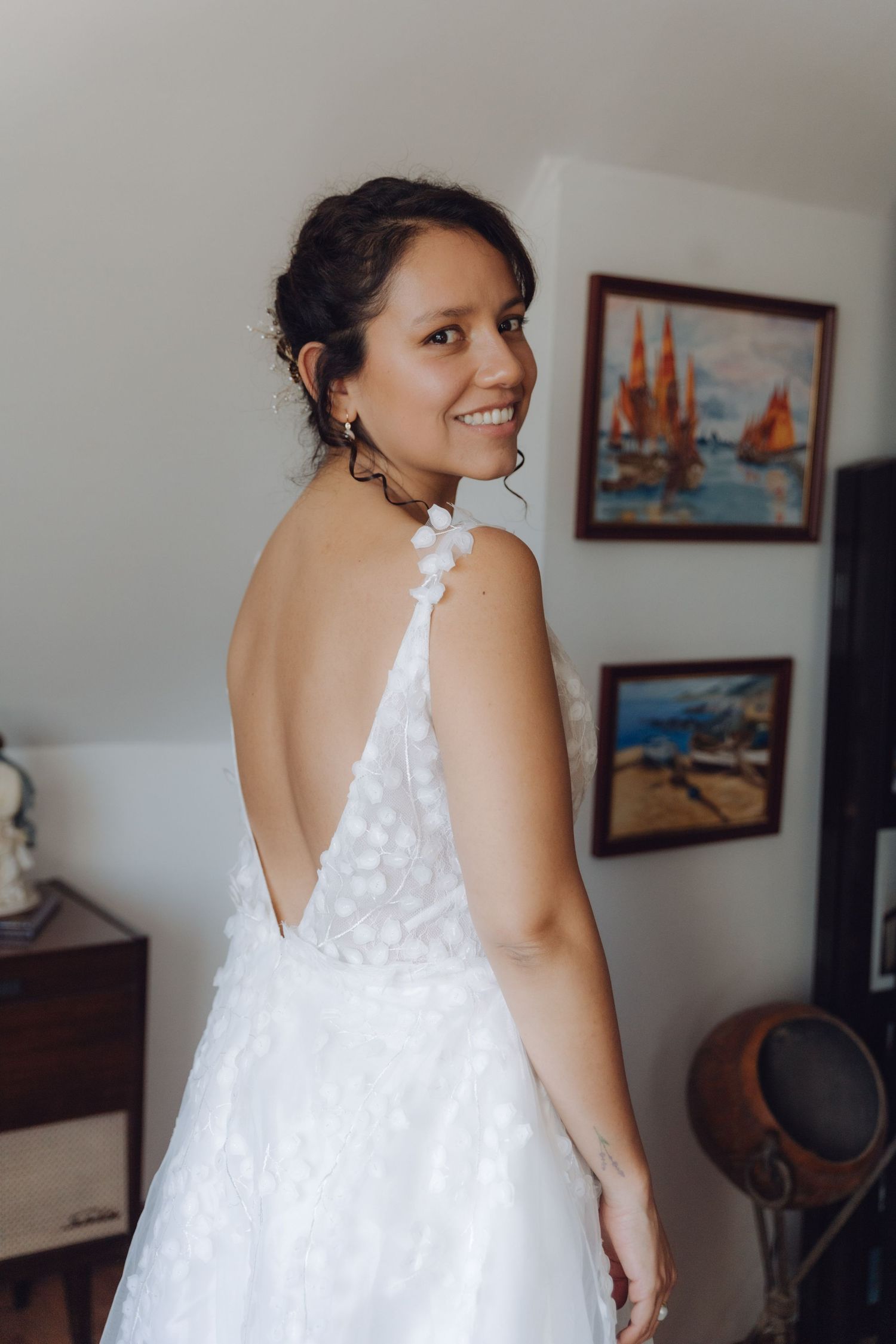 Radiant bride in a backless floral wedding dress smiles over her shoulder near nautical paintings.