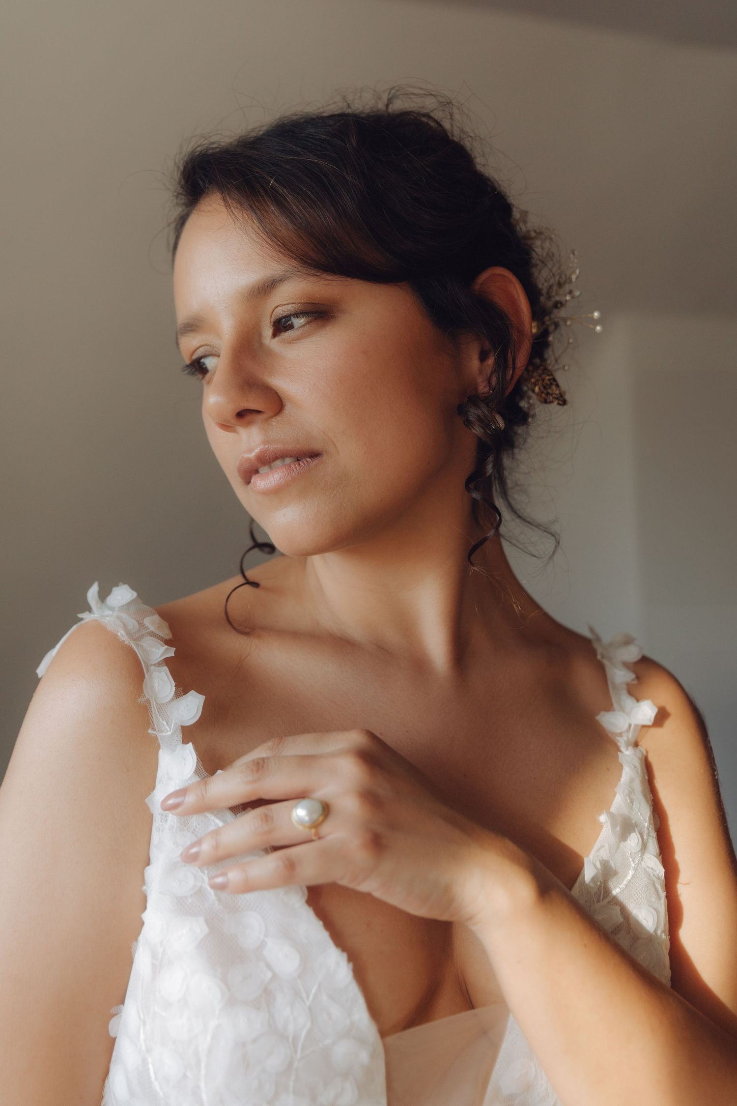 Bride in white floral wedding dress gazes softly while touching her chest, wearing elegant earrings.