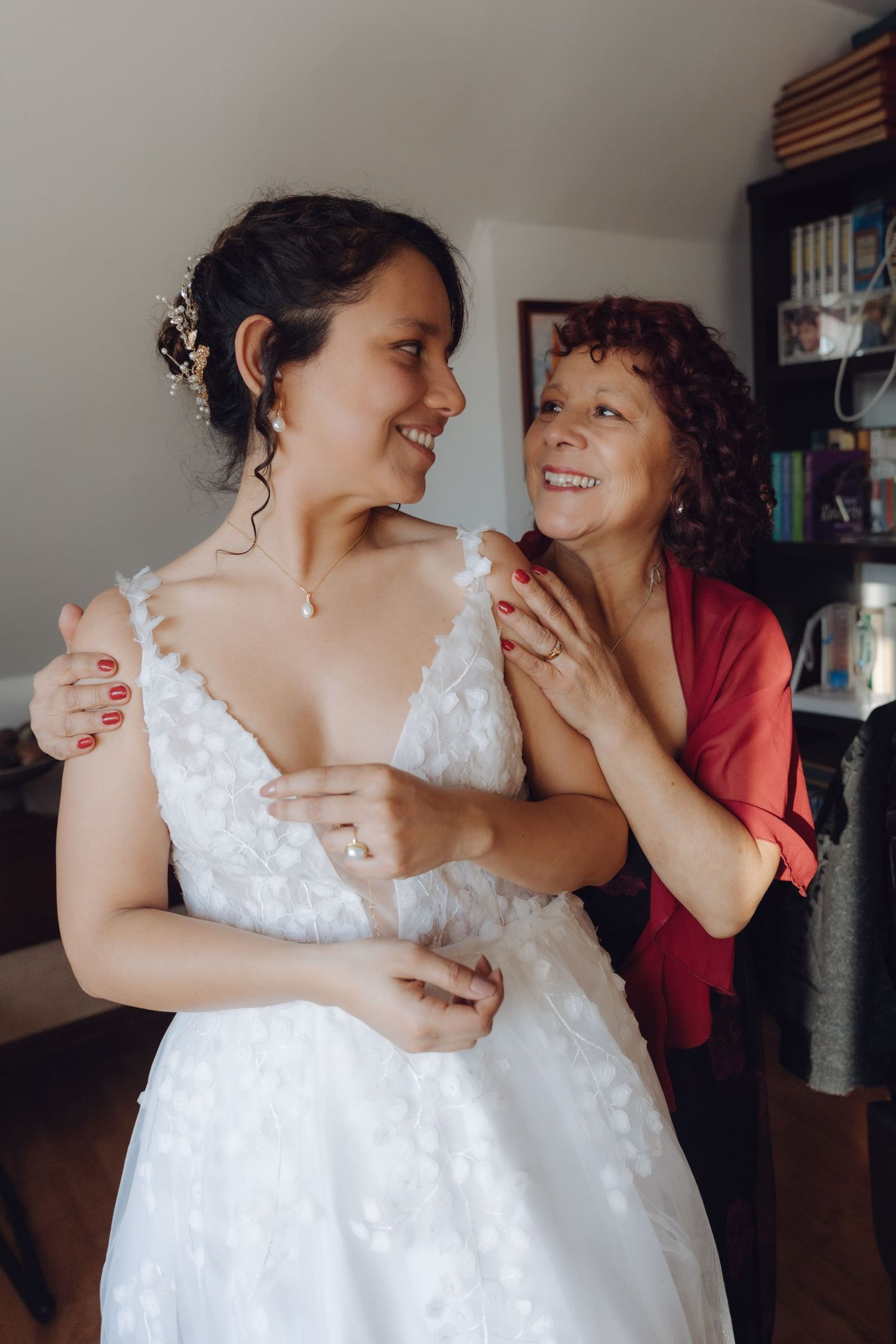 Joyful bride and older woman in red shawl share a happy moment during wedding preparations.