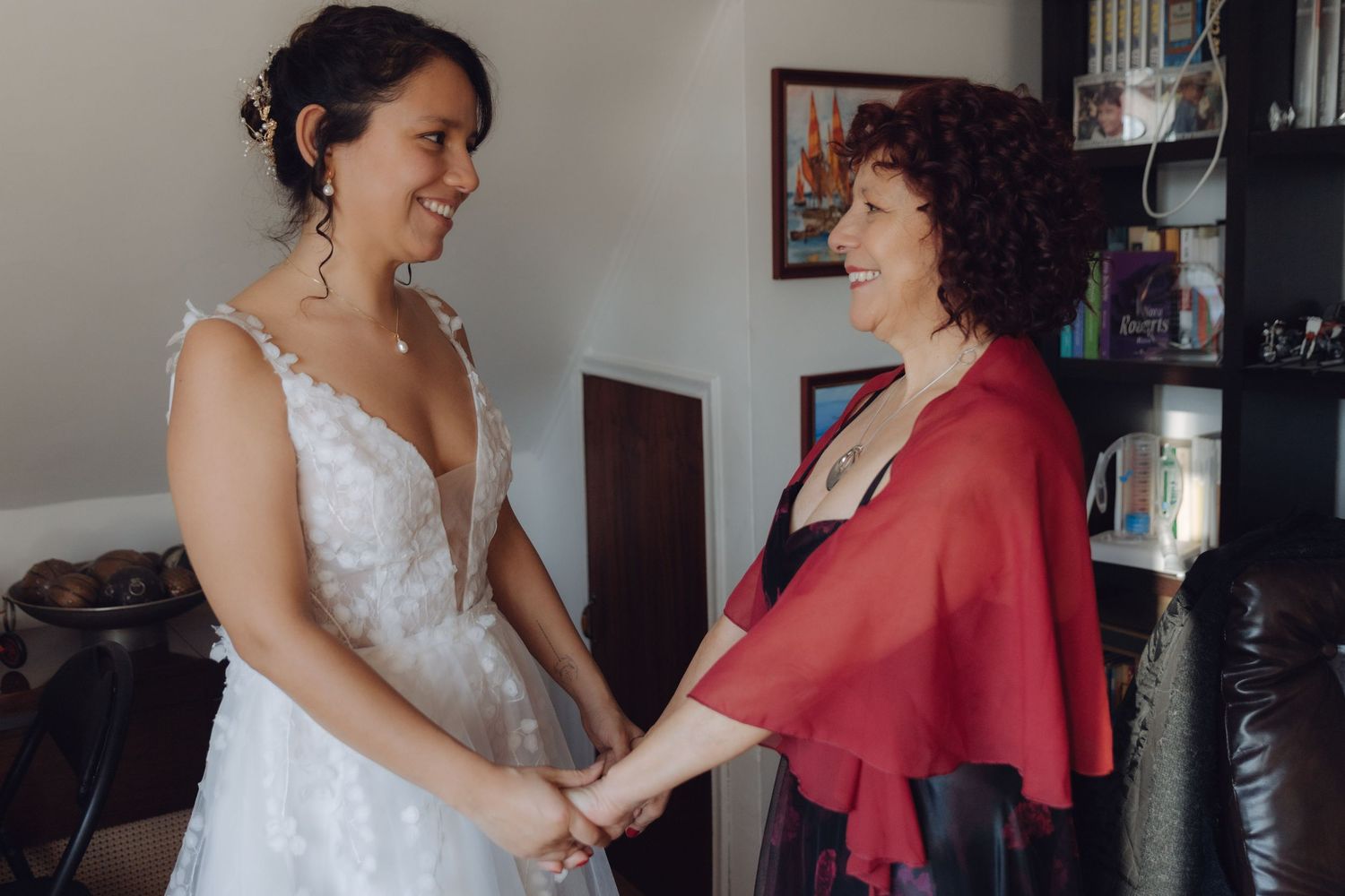 Bride in floral wedding dress holds hands and smiles with older woman in red shawl indoors.