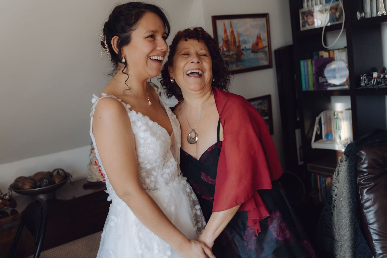 Laughing bride in white wedding dress embraces smiling older woman in red shawl at home.