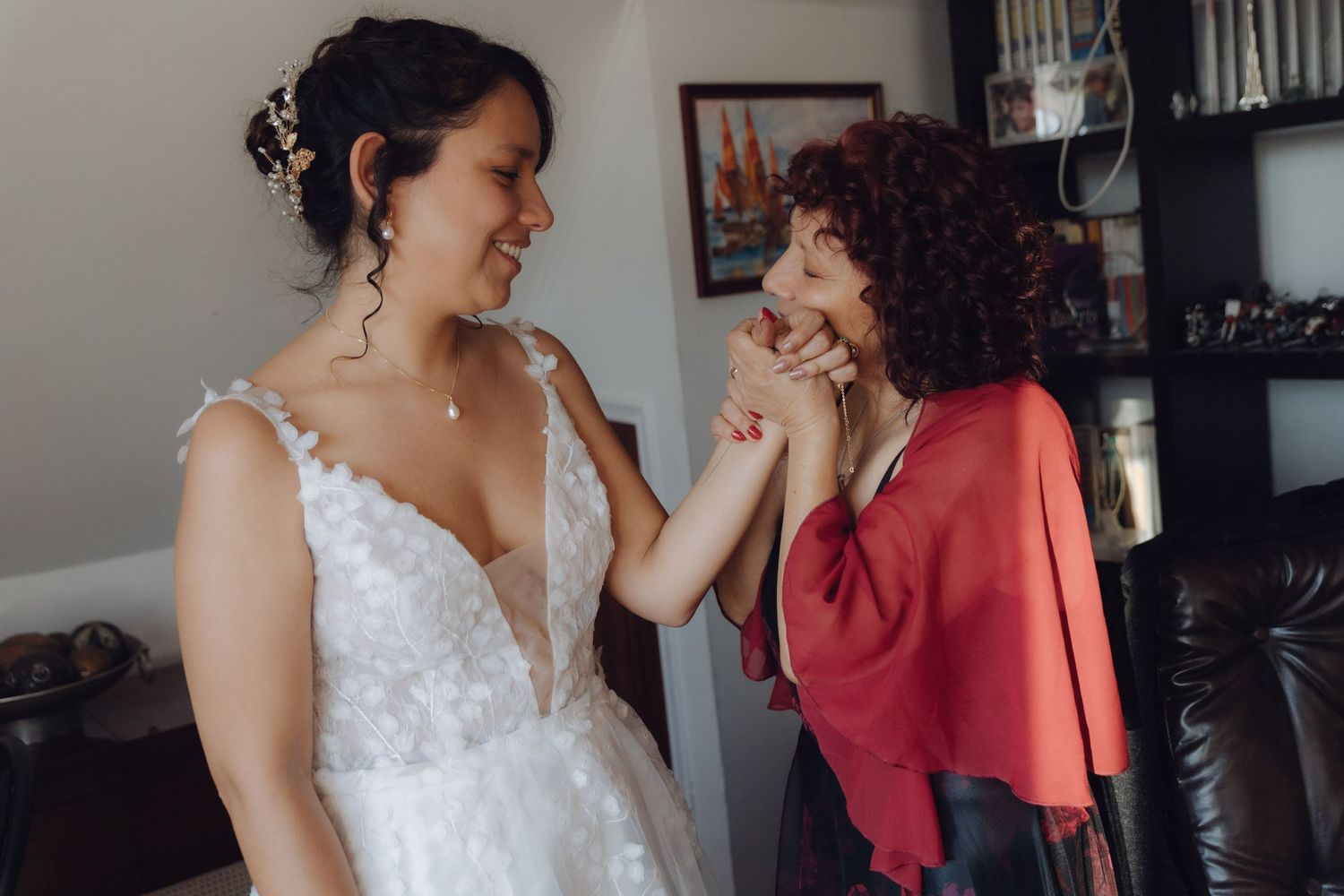 Bride in white floral gown shares a tender moment with older woman in red shawl indoors.