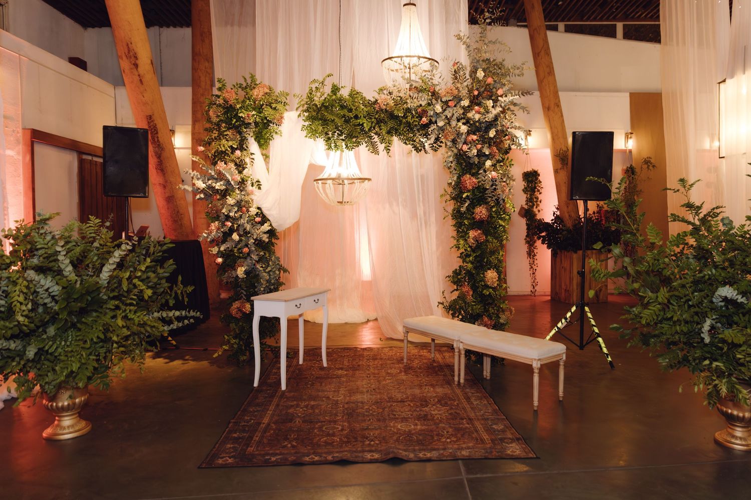 Elegant wedding ceremony arch adorned with lush greenery and pink florals in a warmly lit venue.