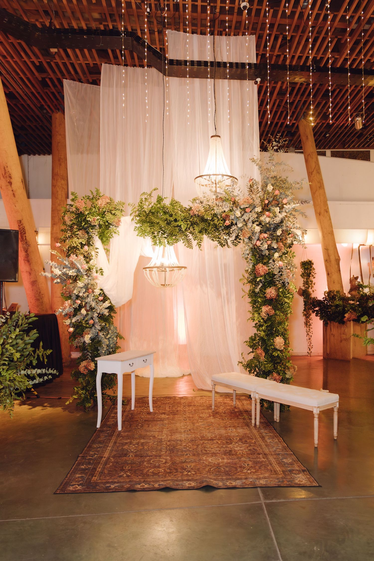 Elegant wedding ceremony backdrop with white draping, floral arch, chandelier, and white bench on vintage rug.