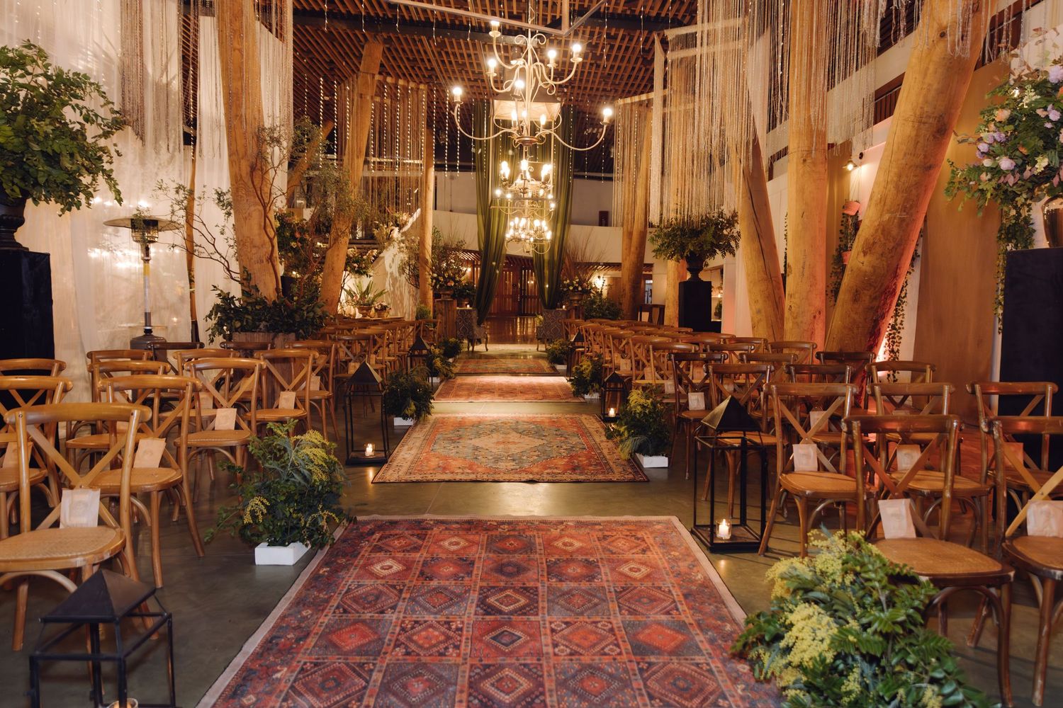 Wedding ceremony aisle with wooden cross-back chairs, layered rugs, candles, and hanging string lights in barn venue.