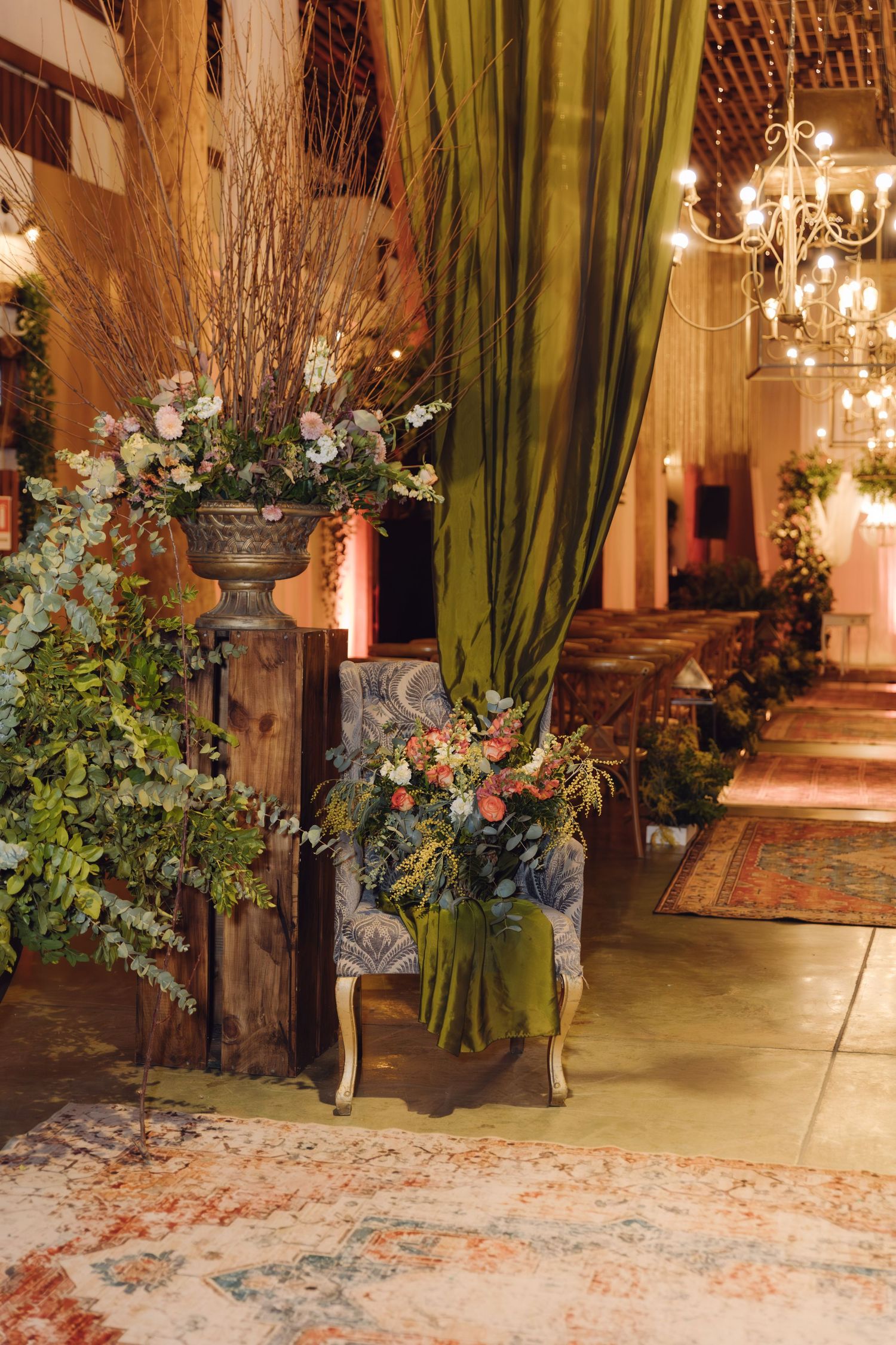 Rustic wedding decor with olive green draping, floral arrangements, and ornate urn on wooden pedestal.