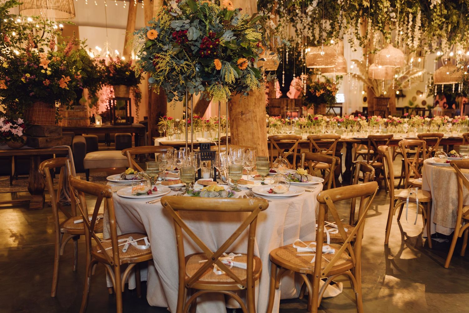 Wedding reception with round tables, cross-back chairs, white linens, and tall greenery centerpieces in warm-lit venue.
