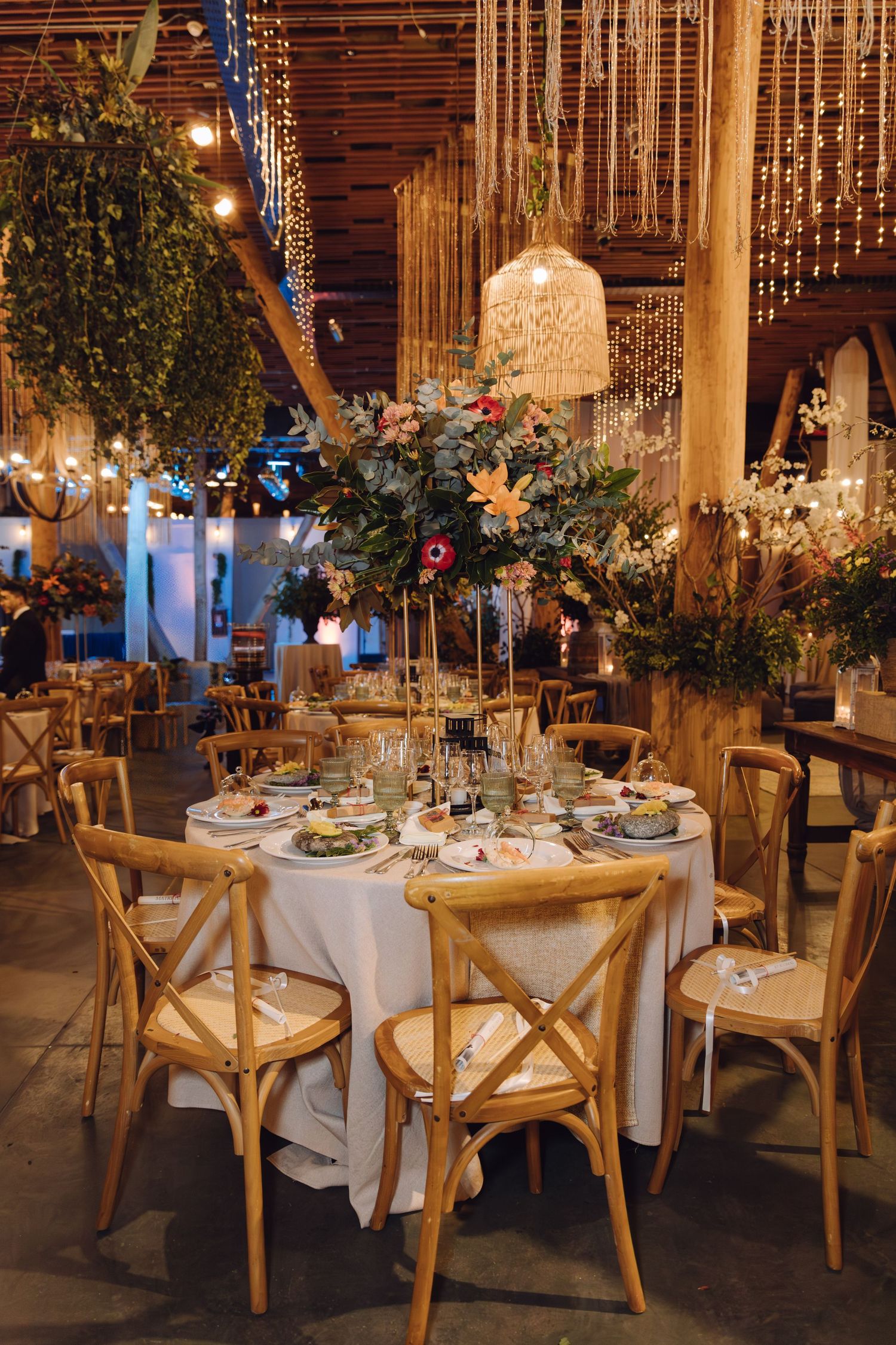 Elegant wedding reception table with cross-back chairs, tall floral centerpiece, and woven pendant lights overhead.