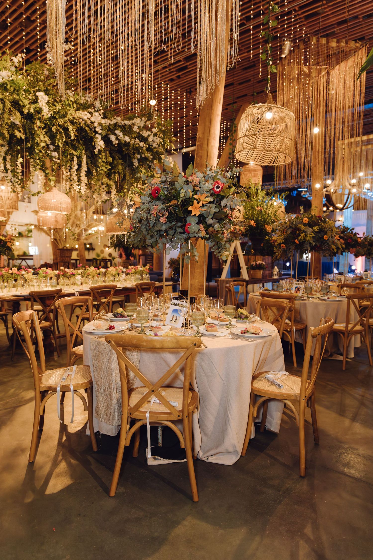 Rustic wedding reception with wooden cross-back chairs, lush floral centerpieces, and woven pendant lights in warm venue.