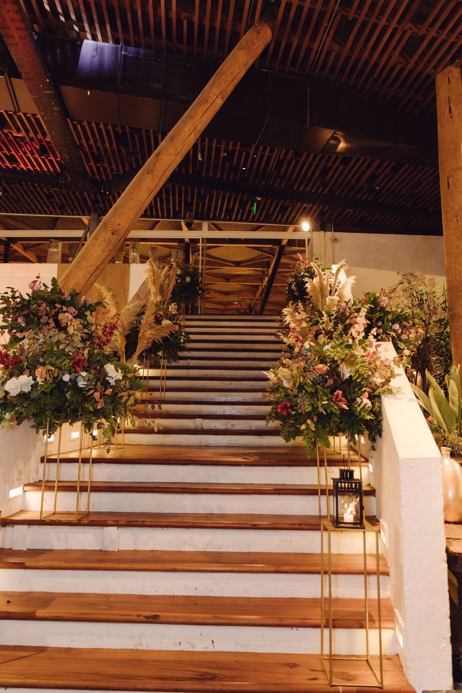 Floral-decorated staircase with gold stands, pink blooms, pampas grass, and lanterns in rustic wedding venue.