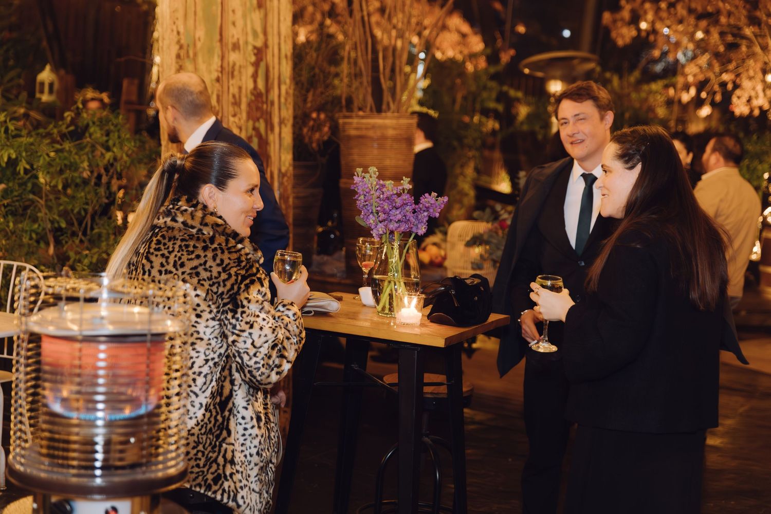 Guests socializing outdoors at evening wedding reception with patio heater and purple floral centerpiece.