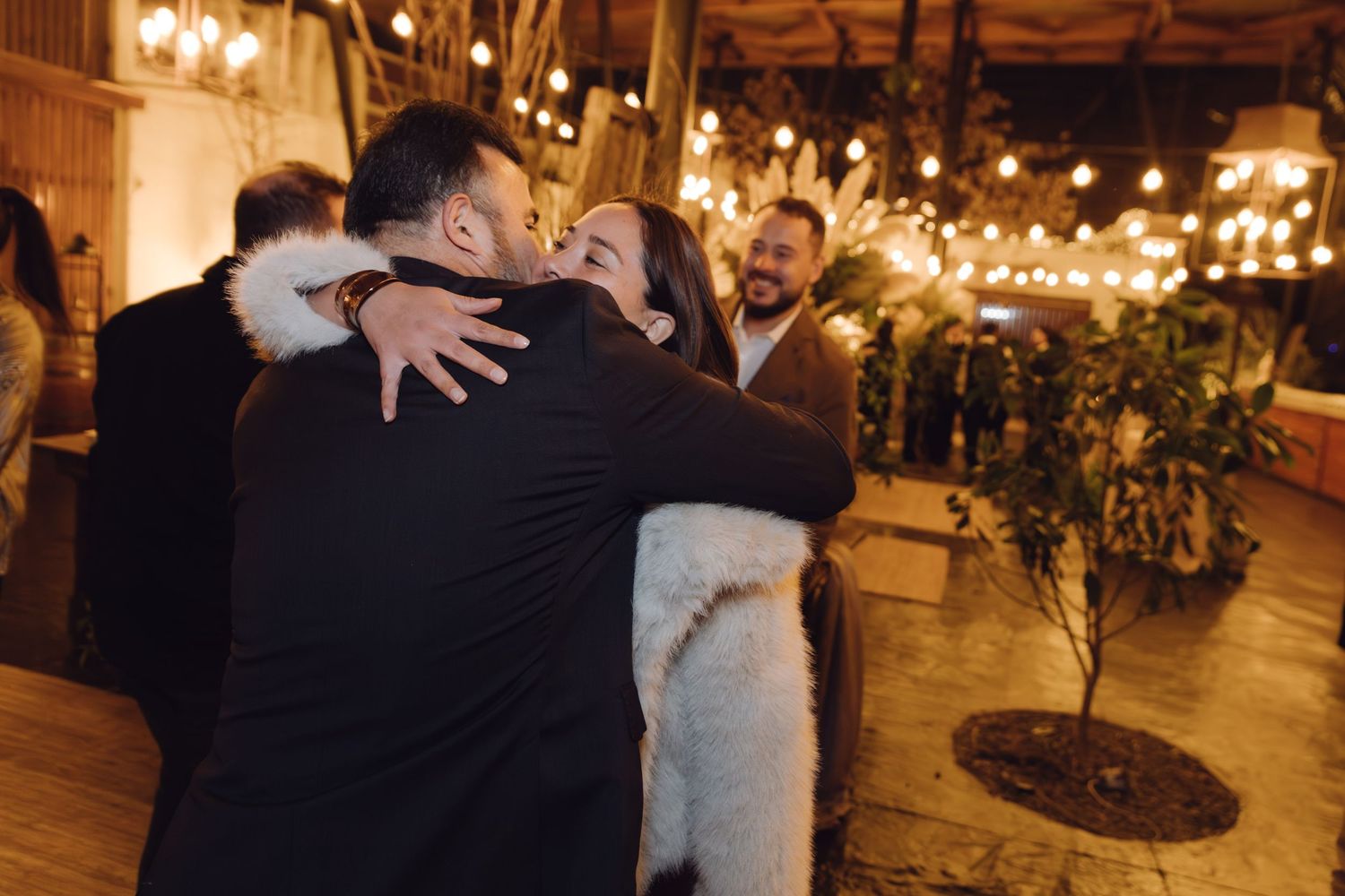 Guests sharing warm embrace at dimly lit wedding reception with string lights and festive atmosphere.