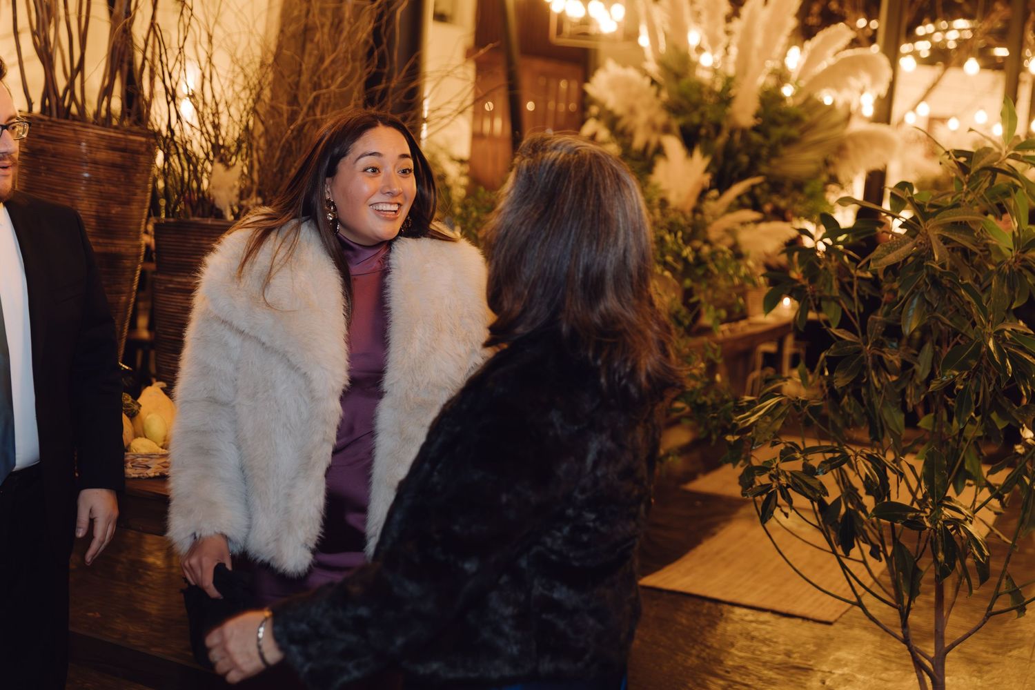 Woman in white fur coat smiling and conversing with another guest at an elegant evening event.