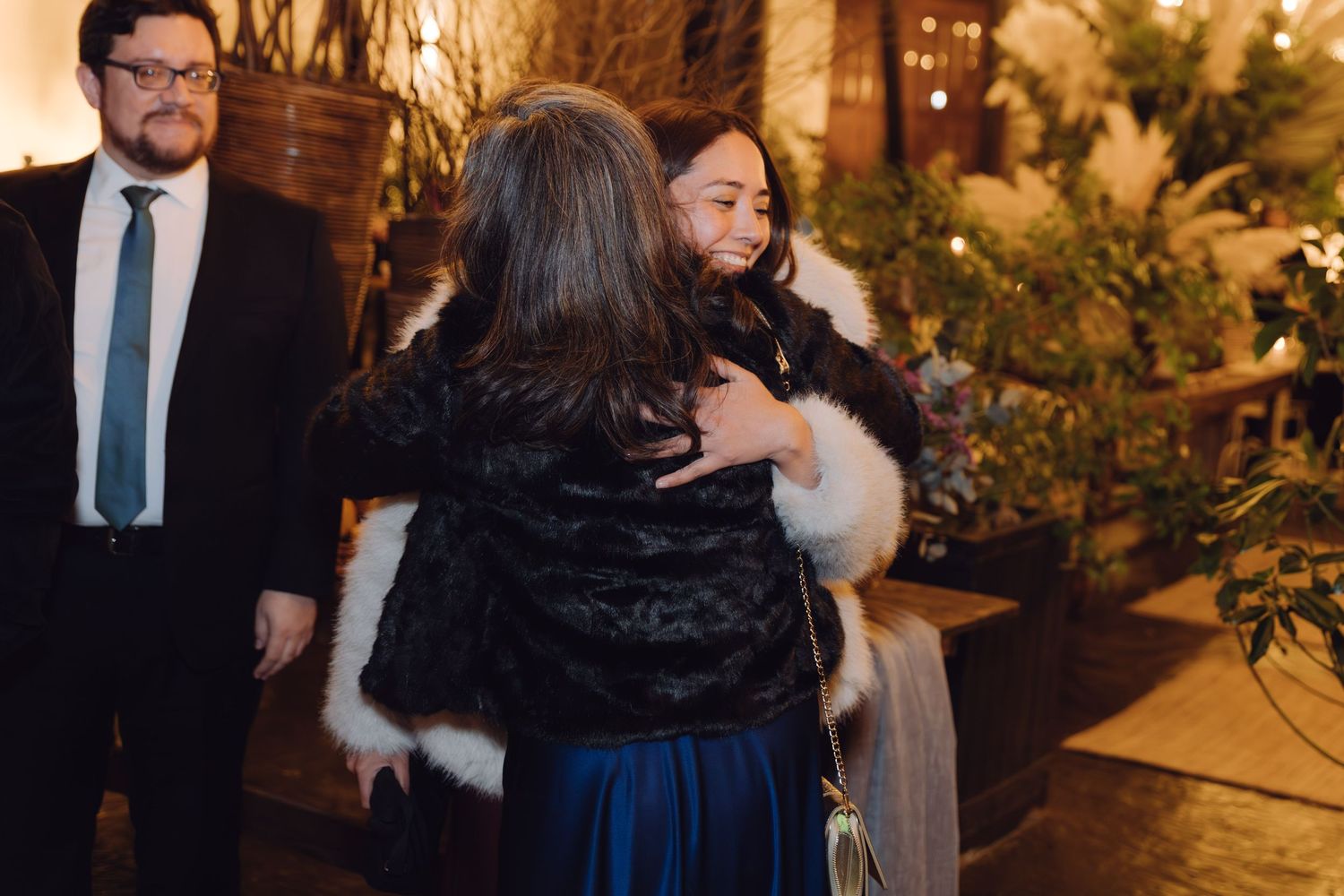 Two women sharing a warm hug at a formal evening event with festive lighting.