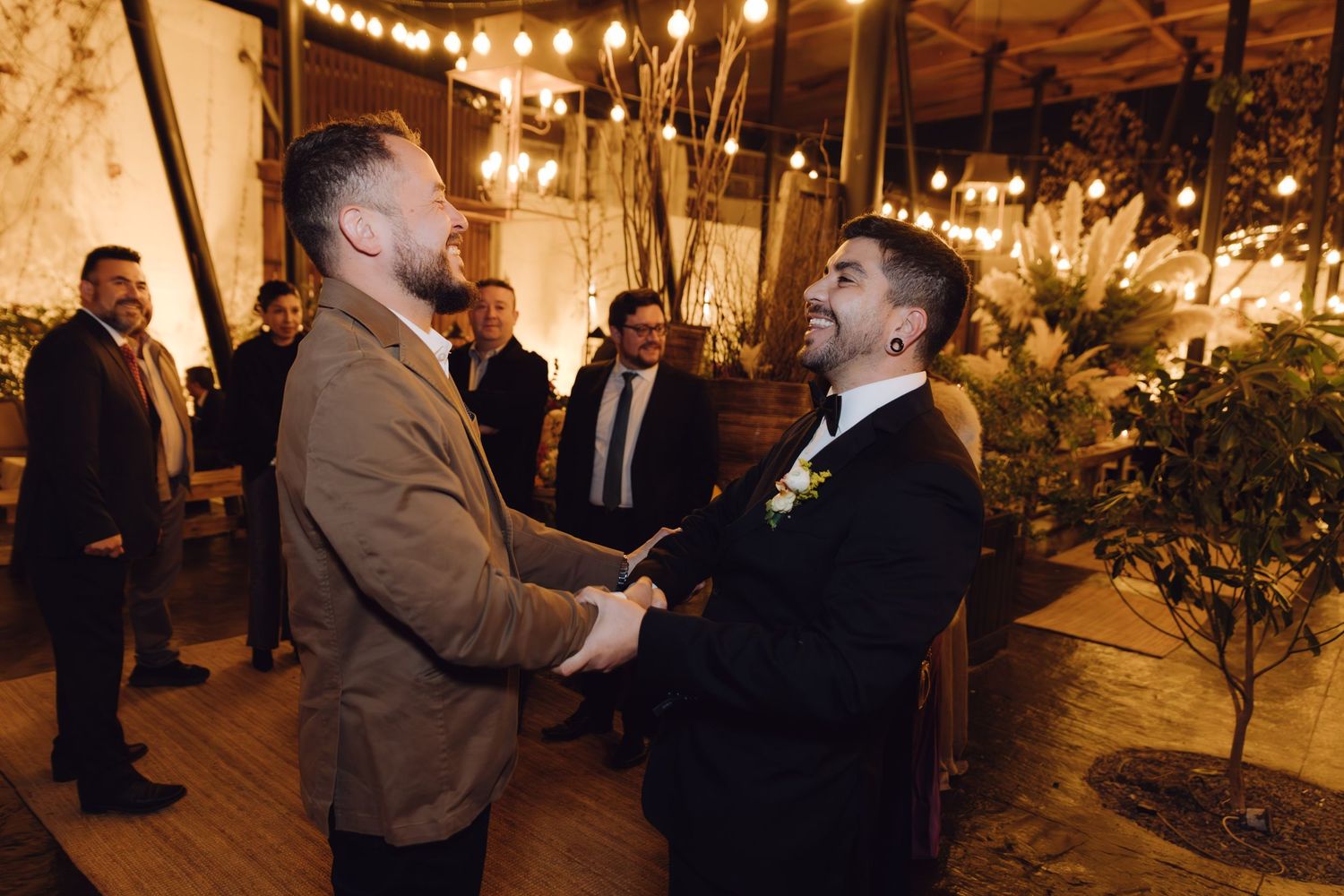 Two men in formal attire shaking hands and smiling at an indoor wedding reception.