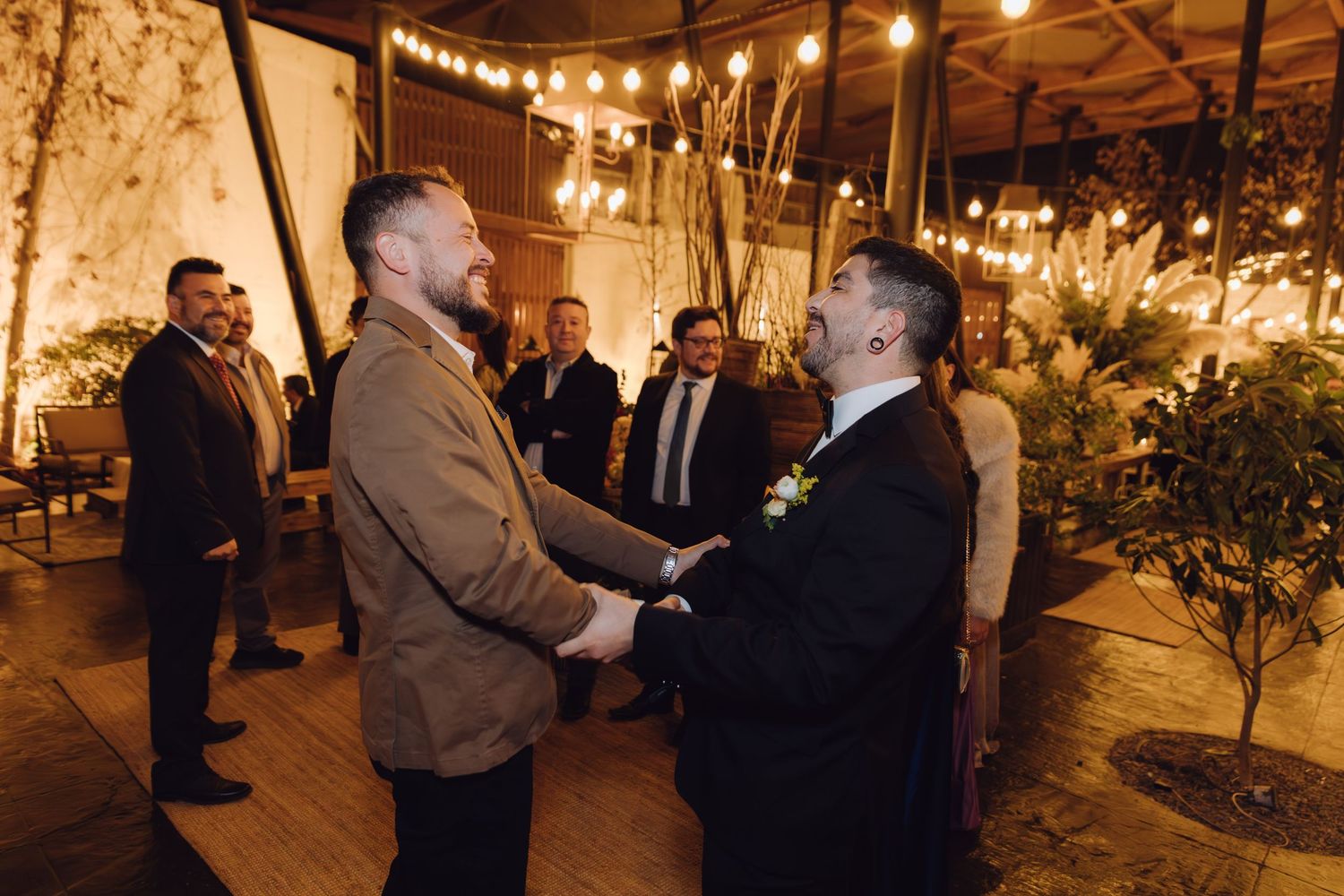 Men in suits greeting each other with a handshake at a warmly lit wedding venue.
