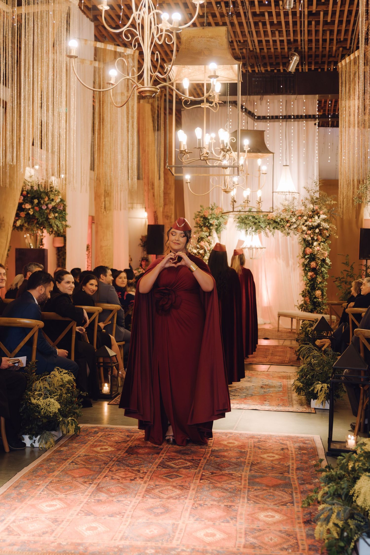 Bridesmaid in burgundy cape dress making heart gesture while walking down wedding aisle.