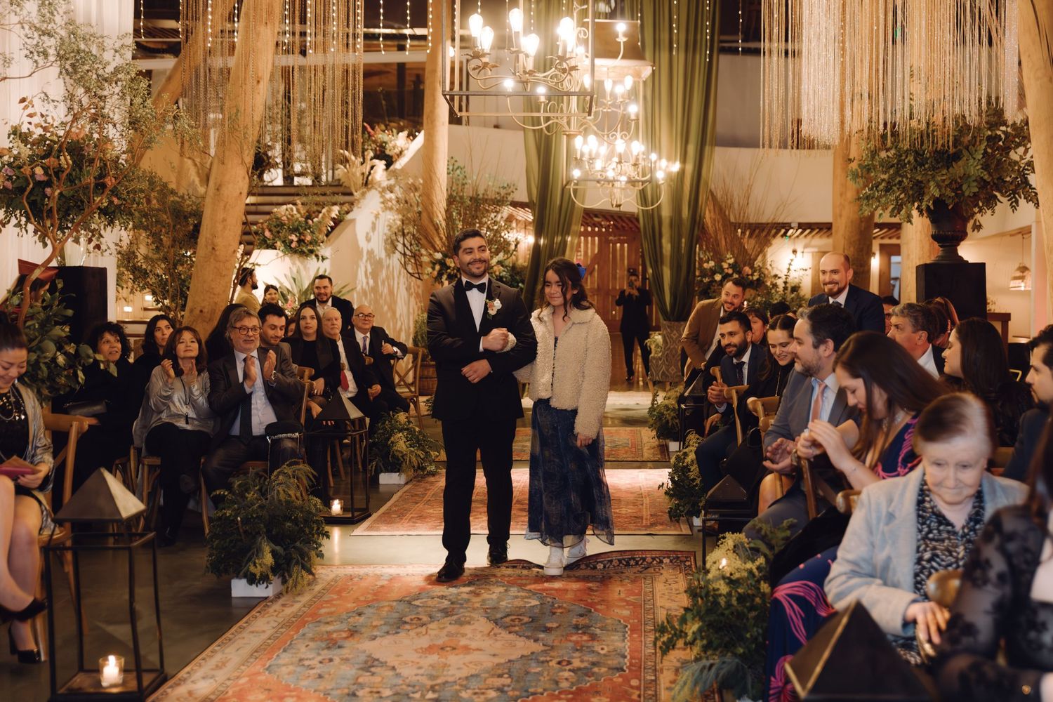 Groom in tuxedo walking down the aisle with a companion at an elegant indoor wedding.