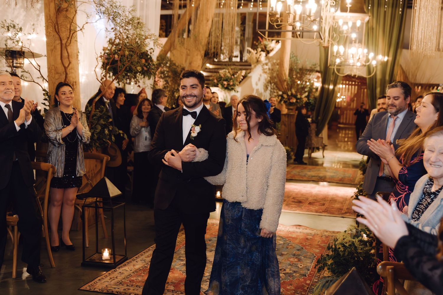 Groom in tuxedo and partner smiling joyfully while guests celebrate at wedding reception.