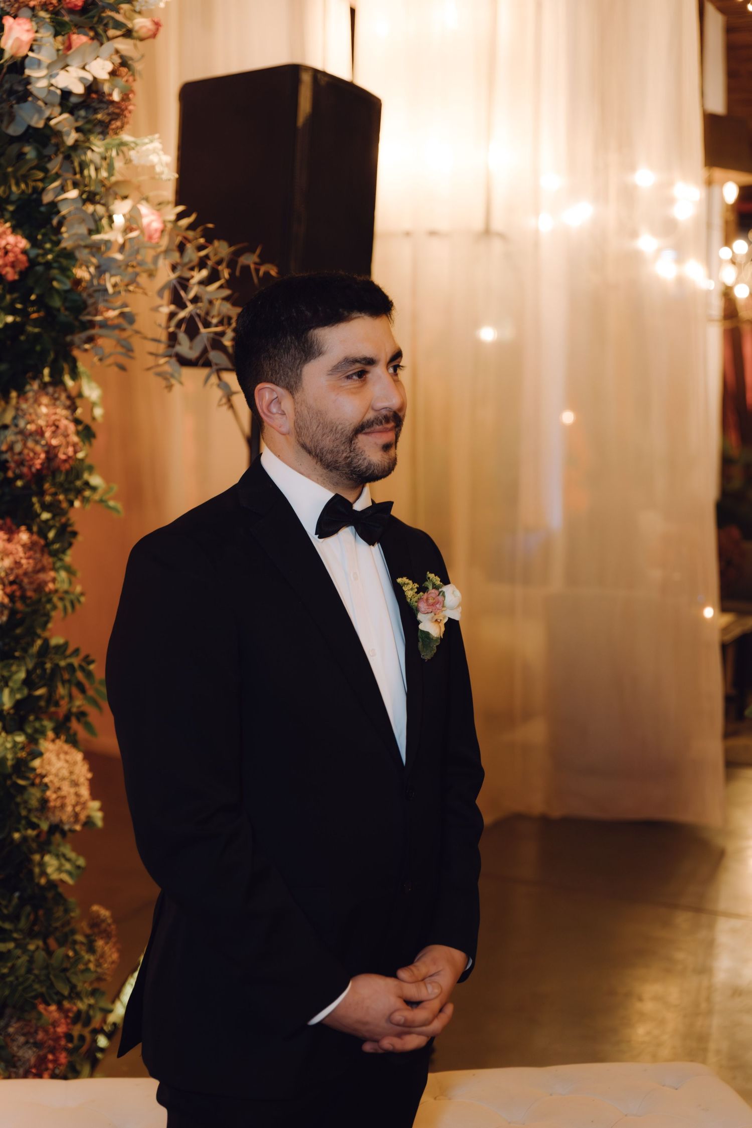 Groom in black tuxedo with boutonniere standing at wedding altar with floral arrangements and string lights.