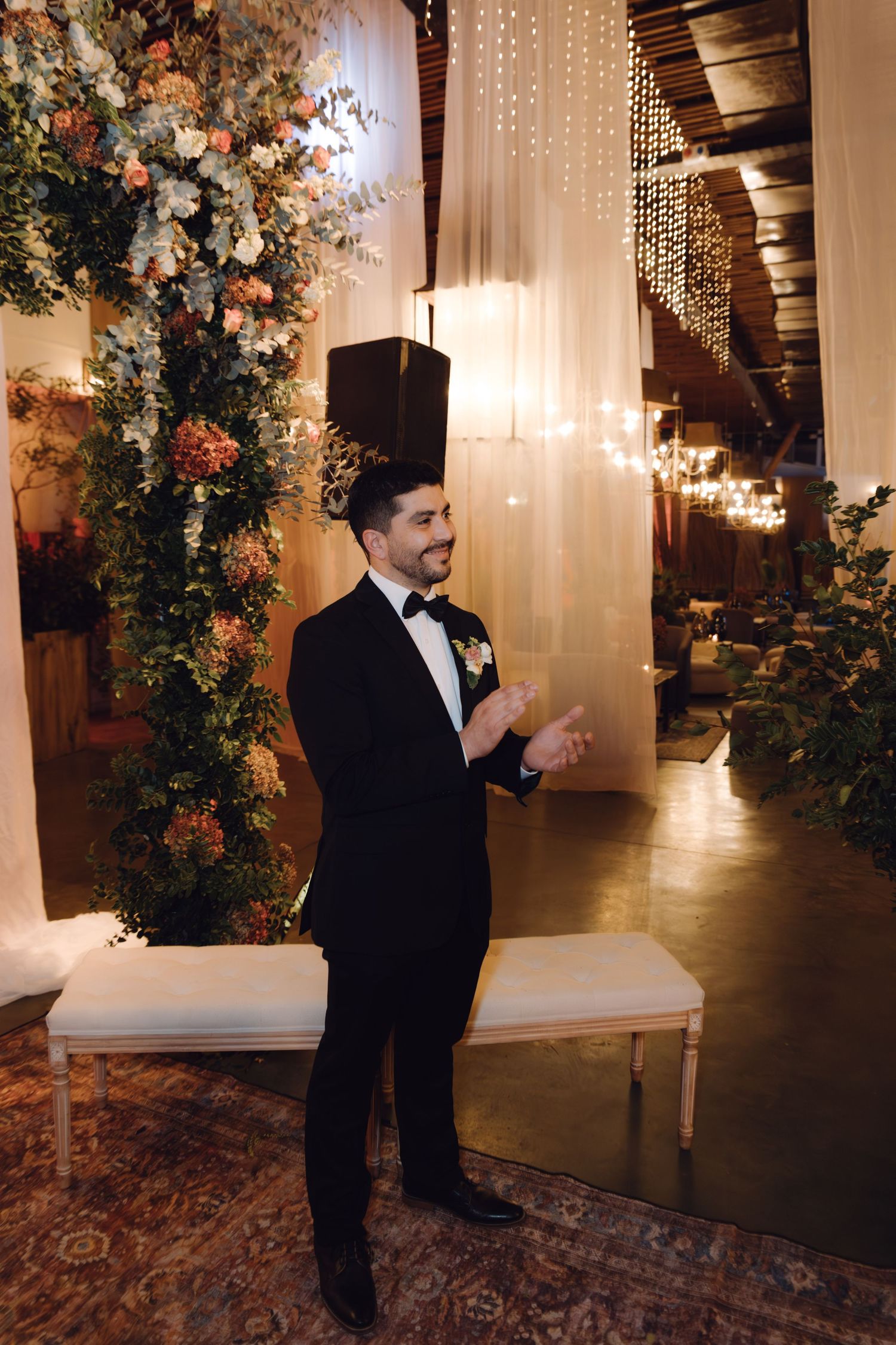 Smiling groom in black tuxedo applauding at elegant wedding venue with floral arch and warm string lights.