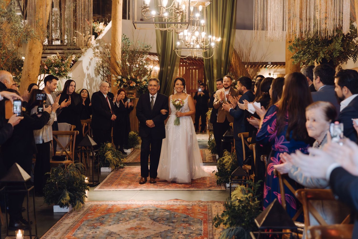 Bride walking down aisle with escort at indoor wedding ceremony with guests applauding and floral decor.