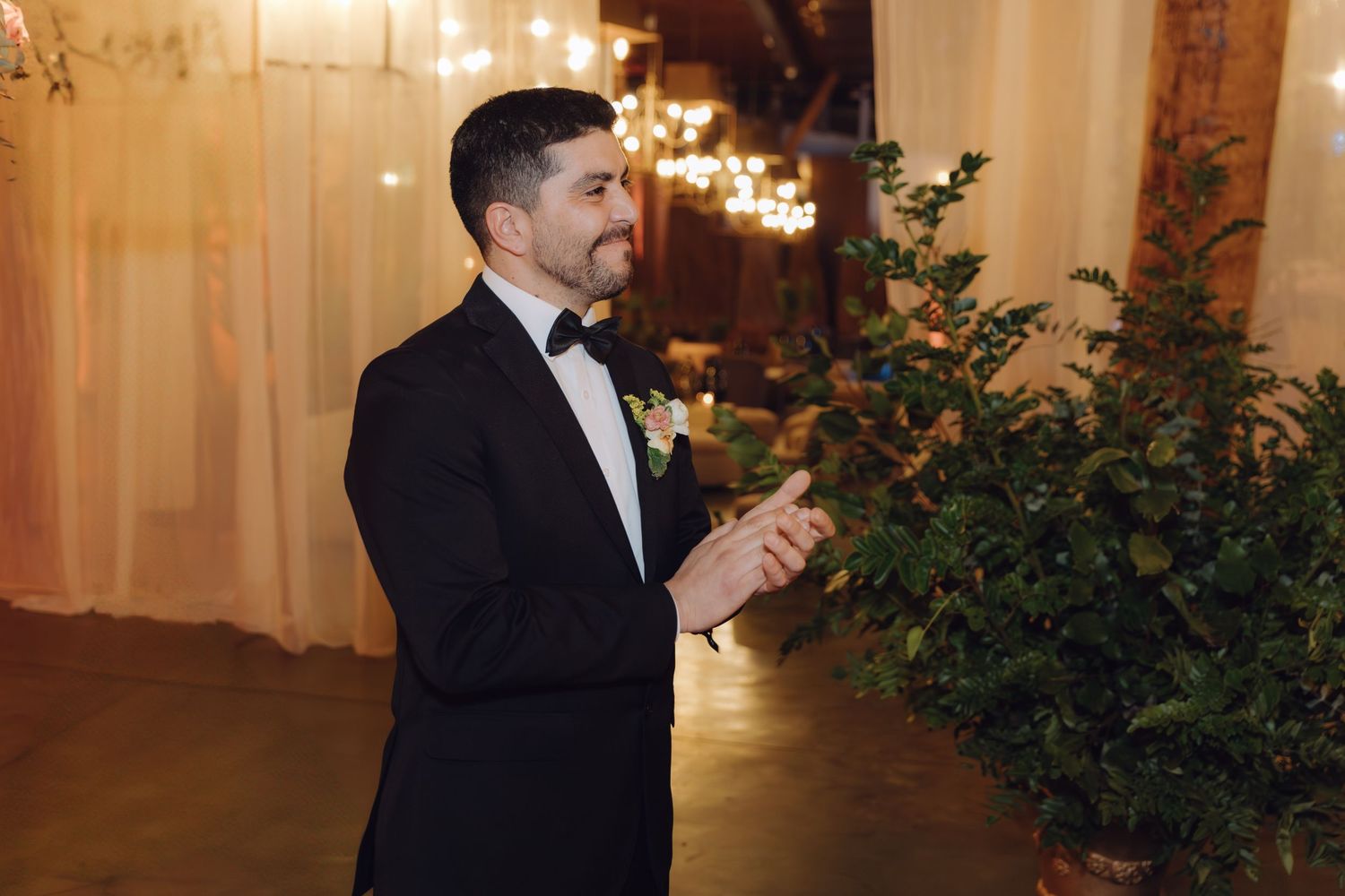 Groom in black tuxedo with boutonniere clapping at warmly lit wedding ceremony venue with greenery.