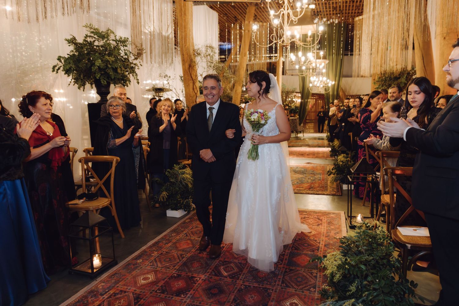 Bride in white gown walking down aisle with escort past applauding guests at elegant indoor wedding.