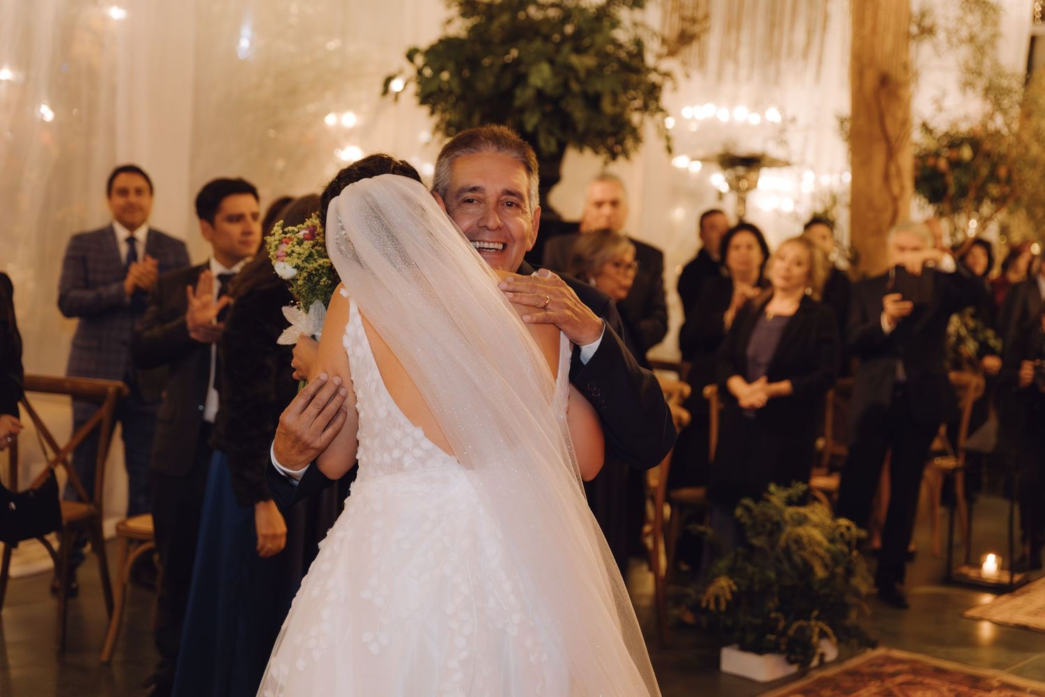 Father of bride sharing emotional hug with daughter in wedding gown at indoor ceremony altar.