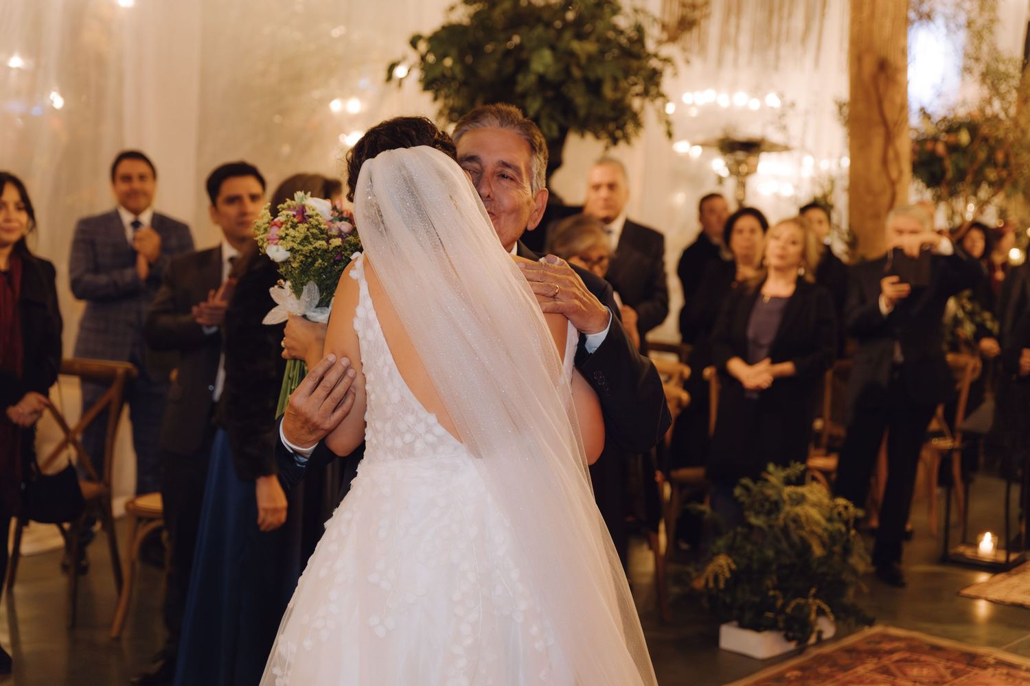 Bride in lace wedding gown embracing escort near altar with seated wedding guests in background.