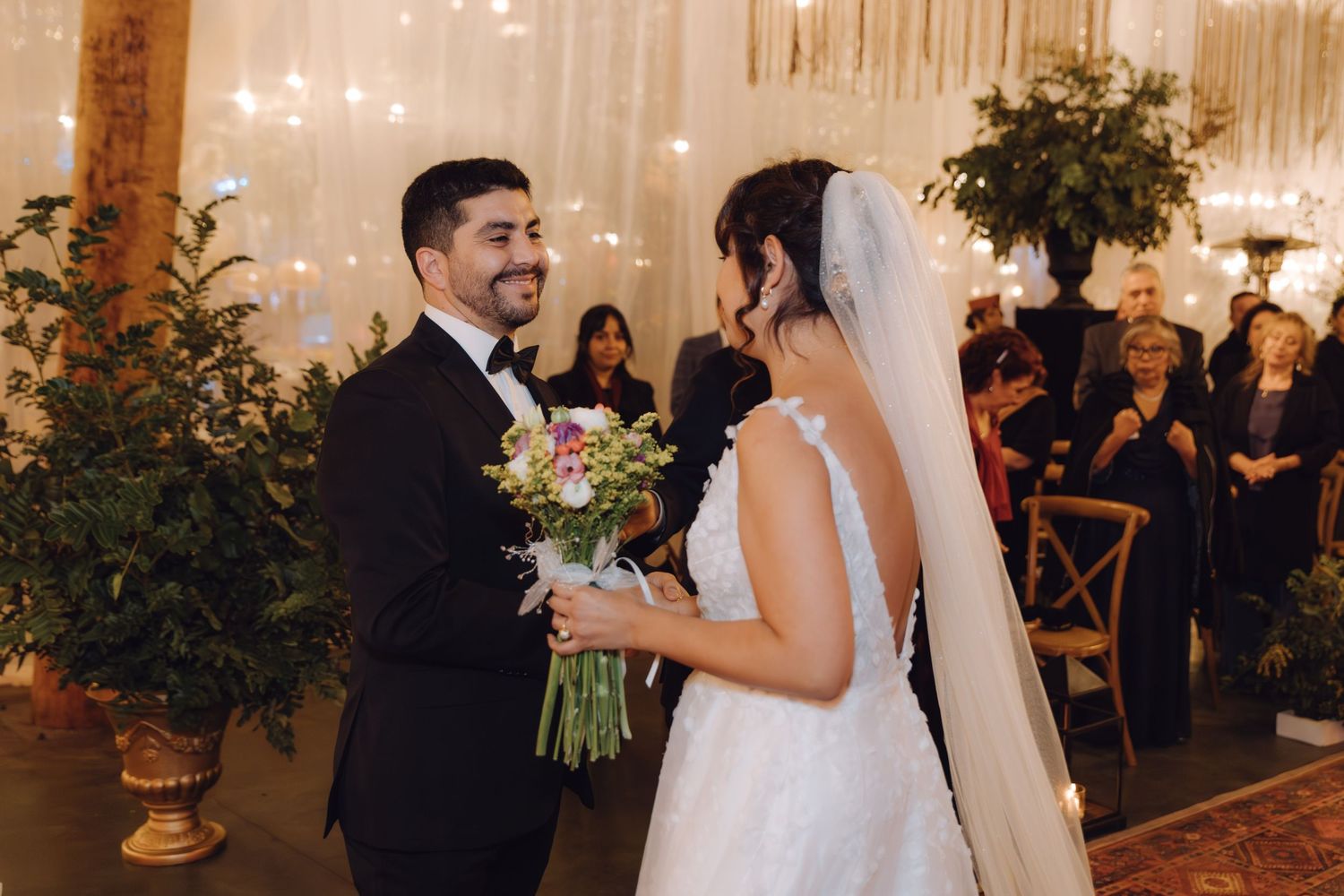 Groom smiling at bride holding bouquet during indoor wedding ceremony with string lights backdrop.