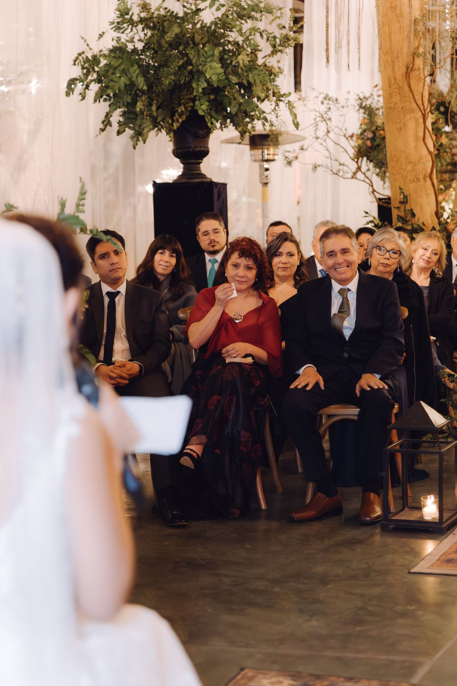 Wedding guests smiling and watching ceremony from seats at elegant indoor venue with greenery decor.