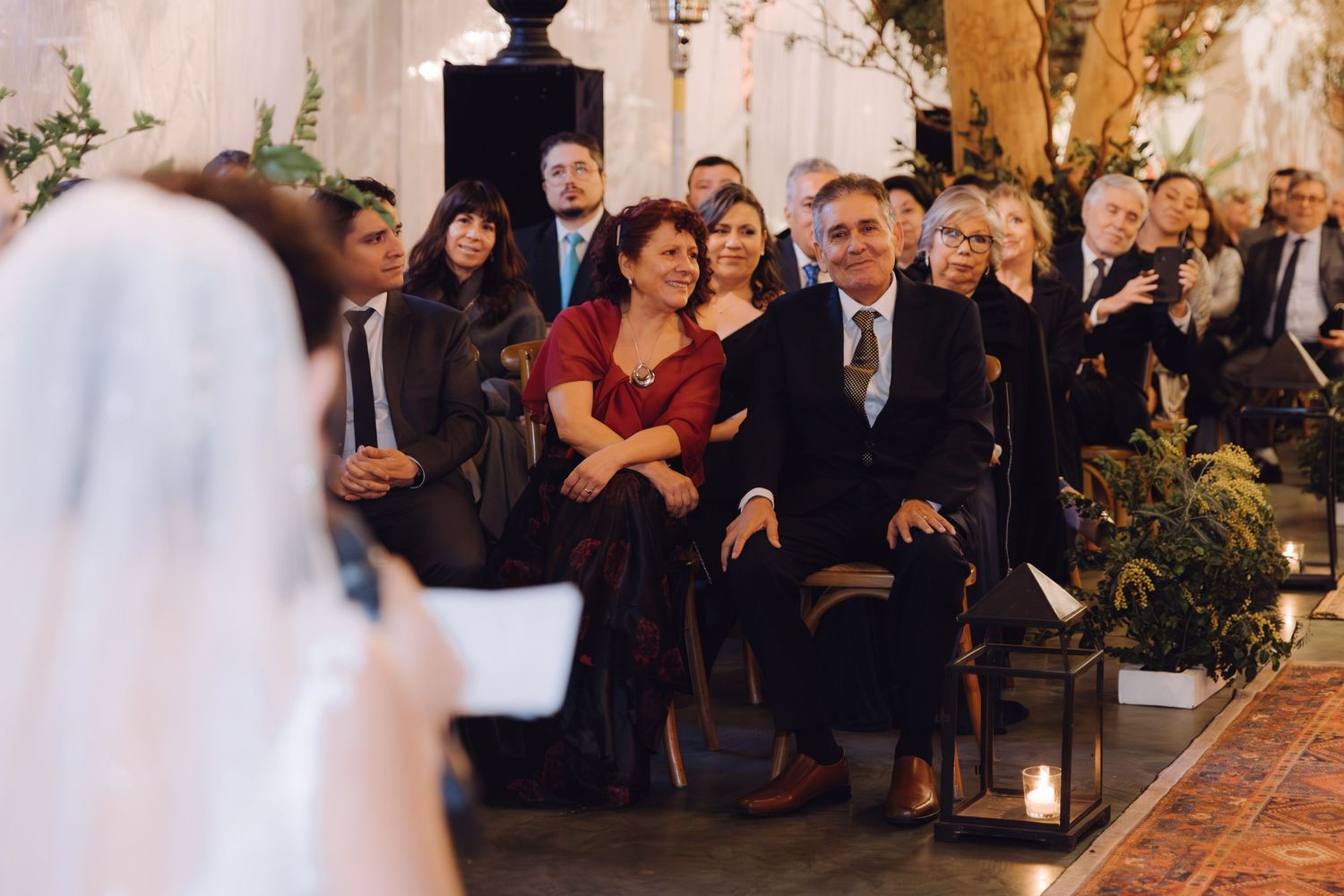 Smiling wedding guests seated at elegant indoor ceremony with candlelit aisle and lush greenery decor.