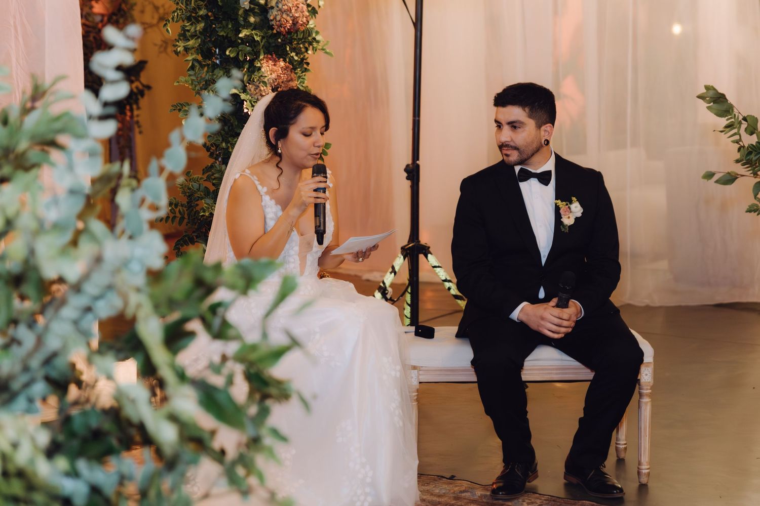 Bride reads vows into microphone while groom listens attentively during elegant indoor wedding ceremony.