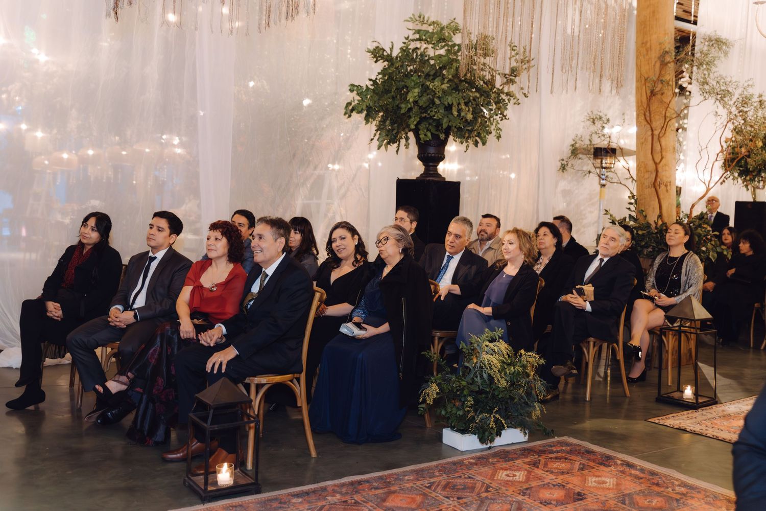 Wedding guests seated in rows watch ceremony in beautifully decorated venue with string lights and greenery.