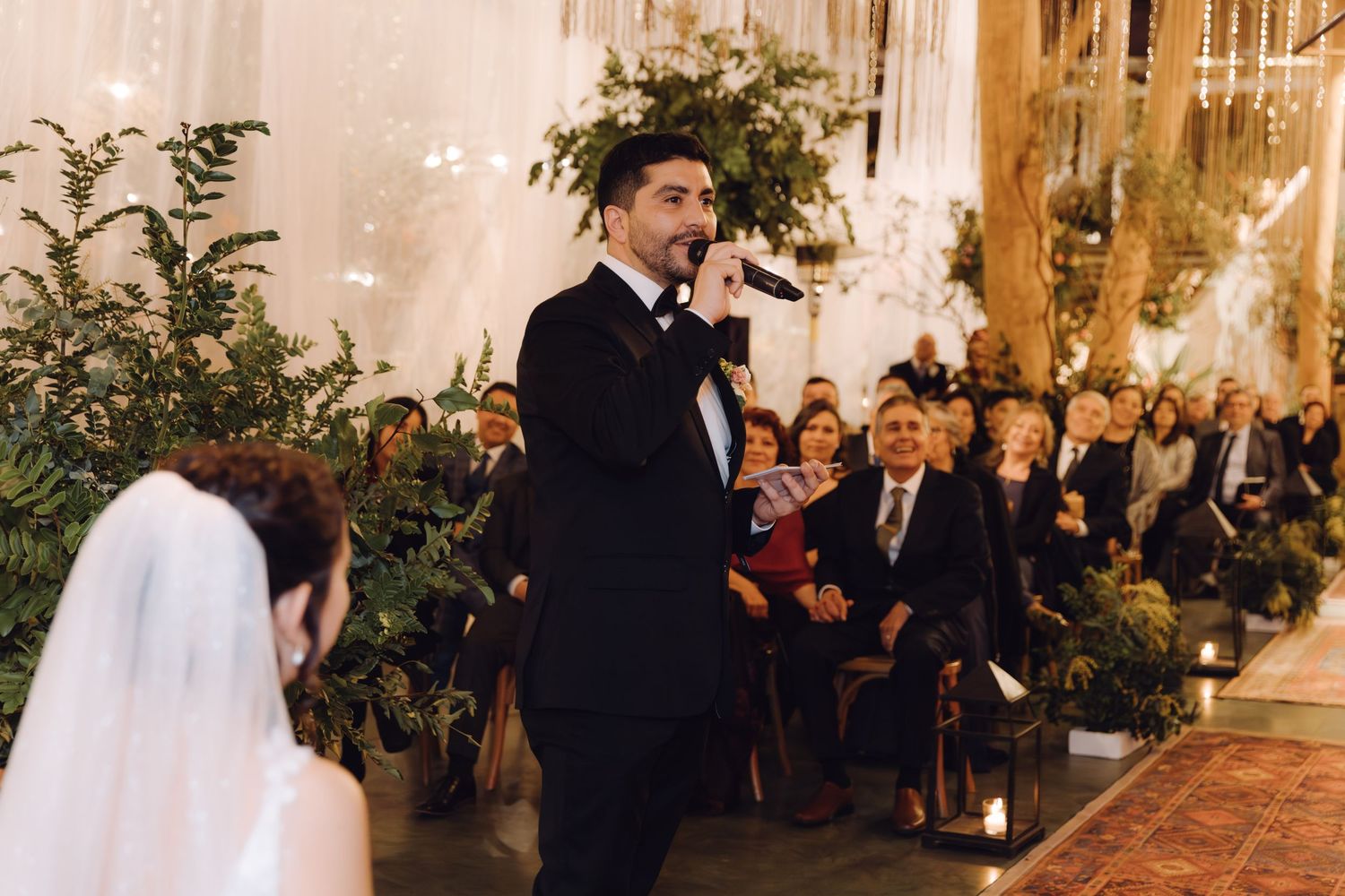 Groom speaks into microphone during wedding ceremony while bride and guests look on in decorated venue.