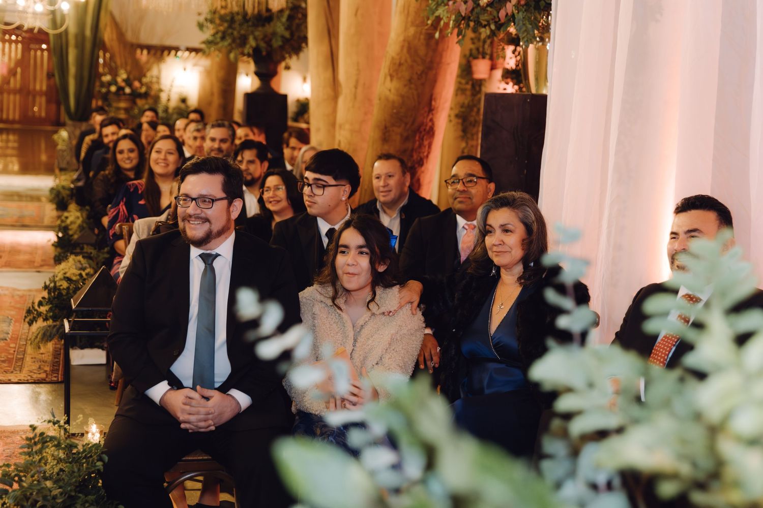 Smiling wedding guests seated among lush greenery decorations during indoor evening wedding ceremony.