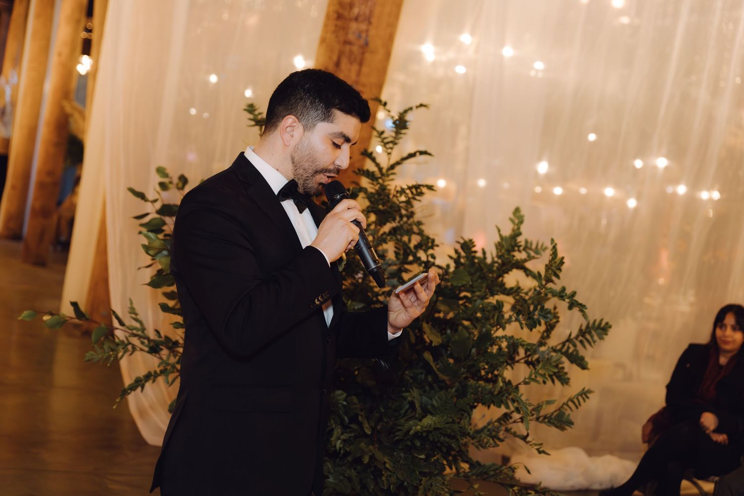 Groom in tuxedo reads from paper while speaking into microphone at elegant wedding ceremony.