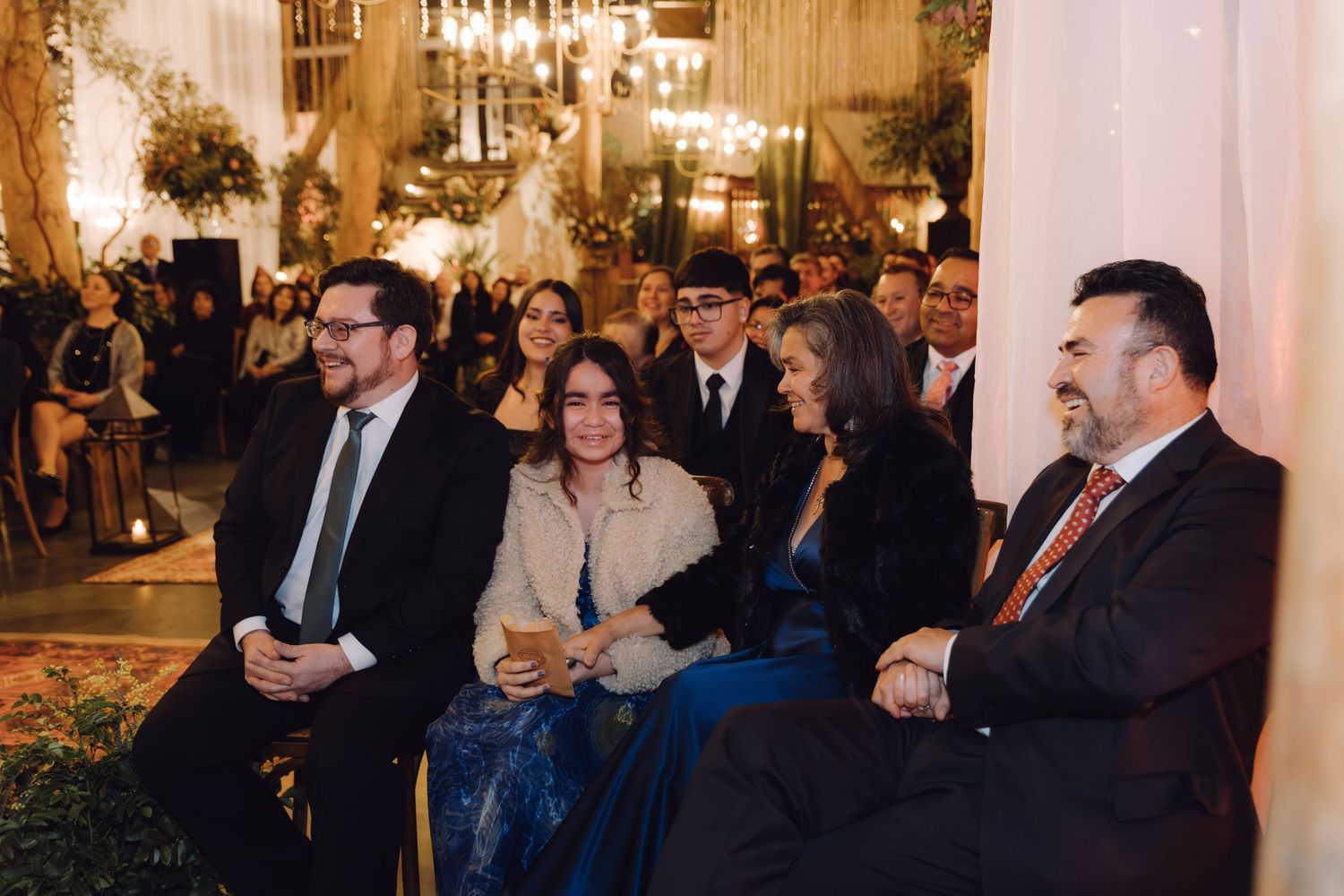 Four elegantly dressed wedding guests smile while seated during indoor wedding ceremony with ornate decor.