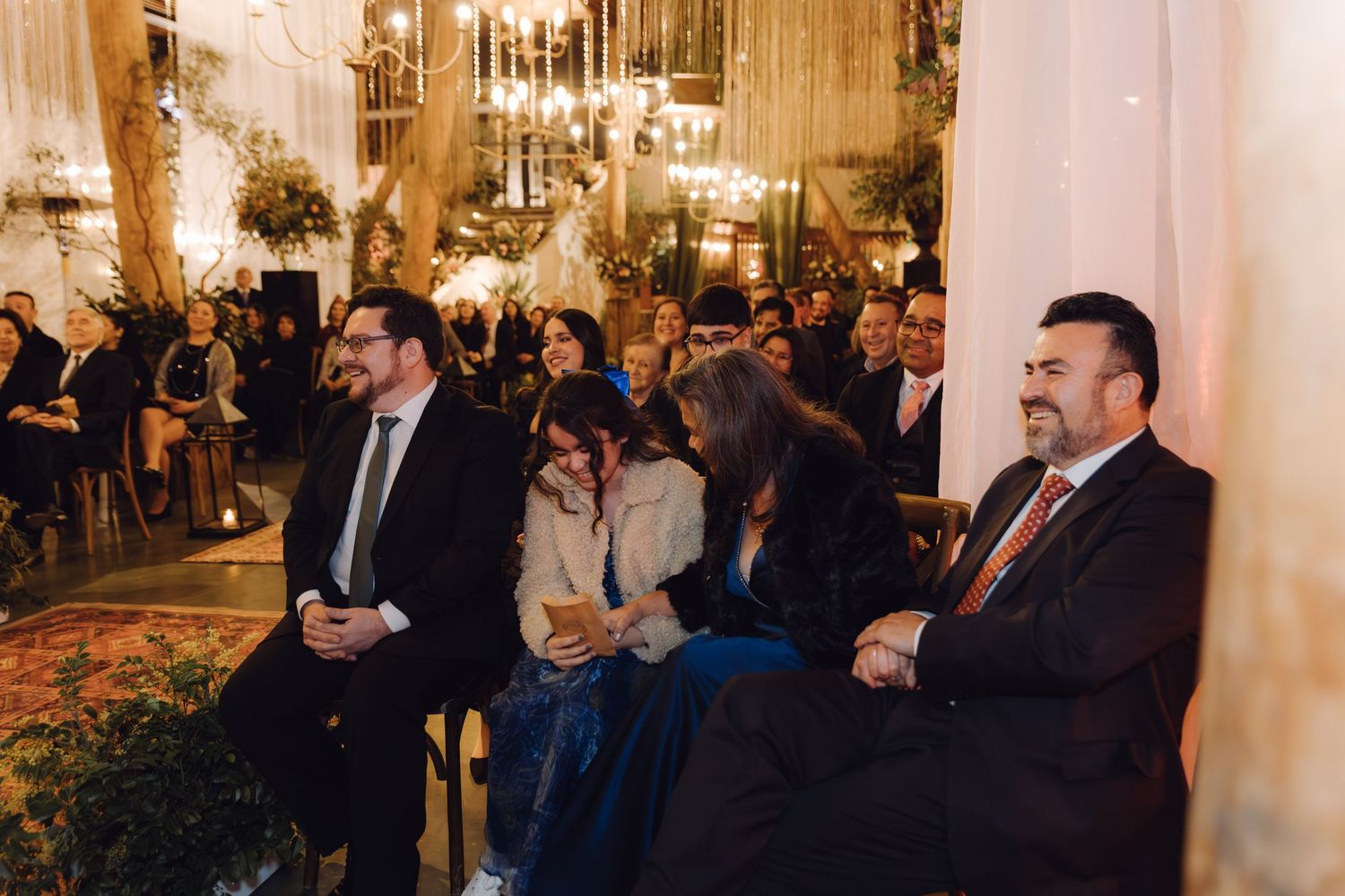 Wedding guests in formal attire smile and watch ceremony in ornately decorated indoor venue.