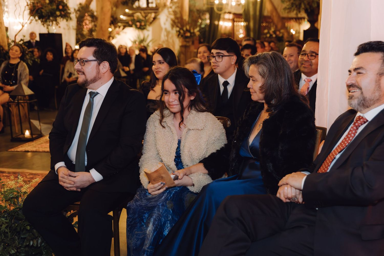 Formally dressed wedding guests seated together smiling during elegant candlelit indoor wedding ceremony.