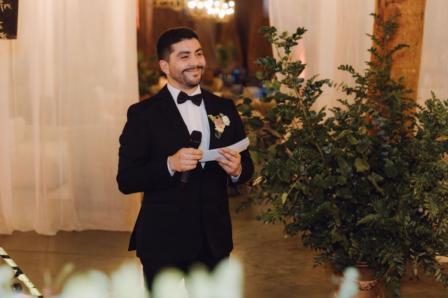 Smiling groom in black tuxedo holds paper and microphone during elegant indoor wedding ceremony.