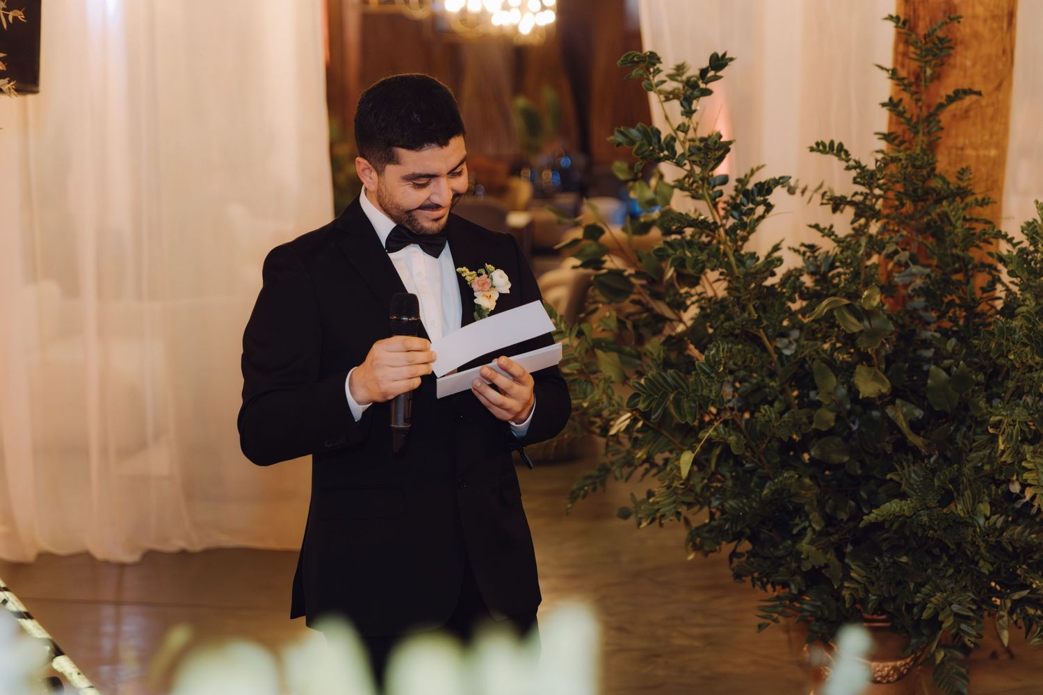 Groom in black tuxedo smiles while reading from paper during wedding ceremony beside greenery.