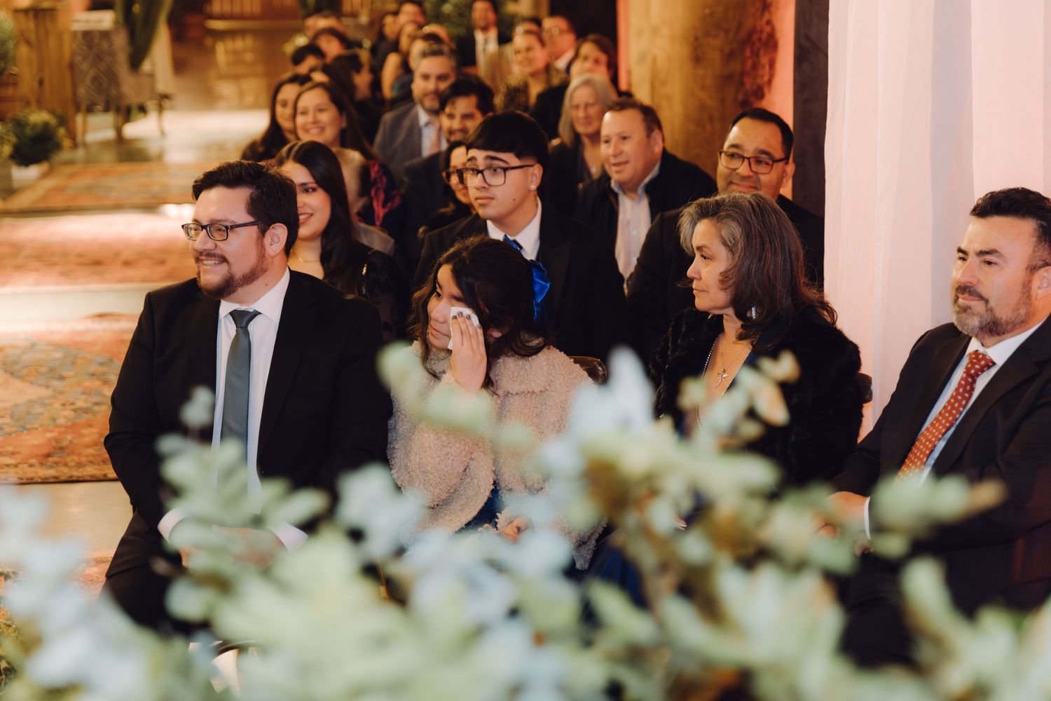 Wedding guests seated and standing during an indoor ceremony, watching attentively with floral decorations in the foreground.