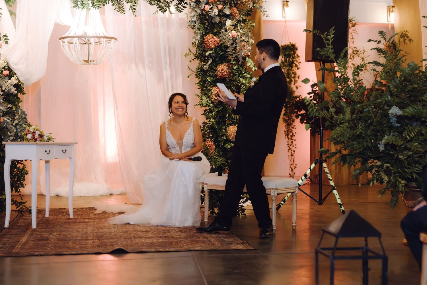 Bride in white gown sits as groom reads vows at elegant indoor wedding ceremony with floral arch backdrop.
