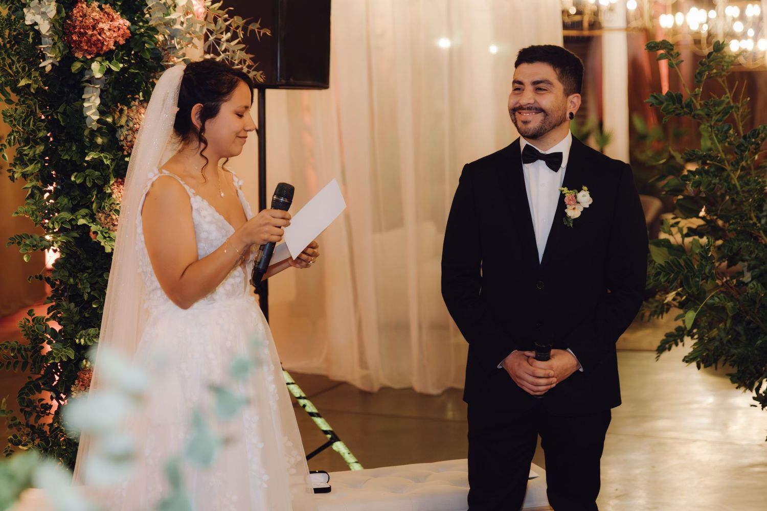 Smiling bride reads vows into microphone while groom in black tuxedo listens at decorated wedding ceremony.
